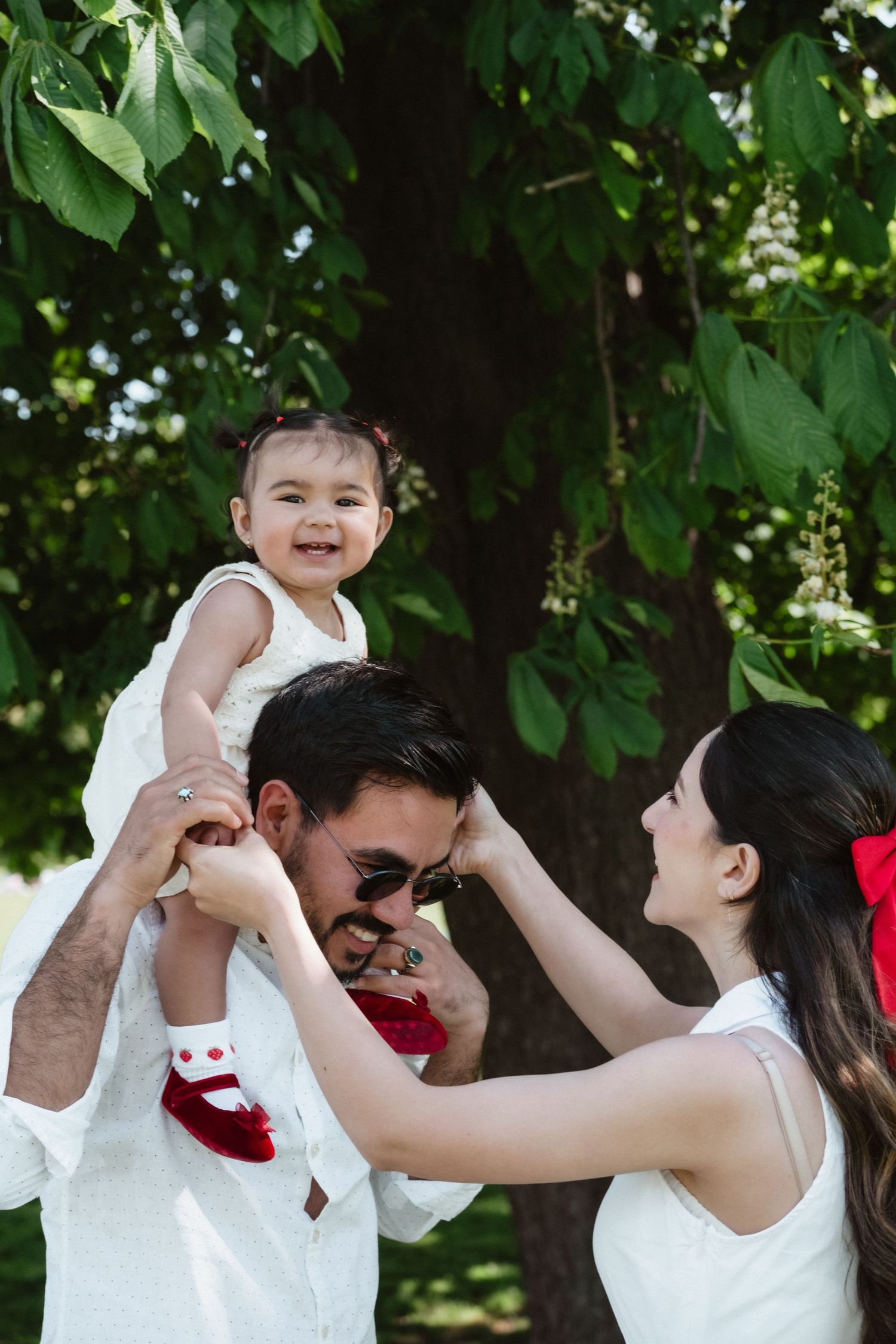 Happy family outdoors, father lifting smiling baby girl on shoulders while mother looks on under green tree.
