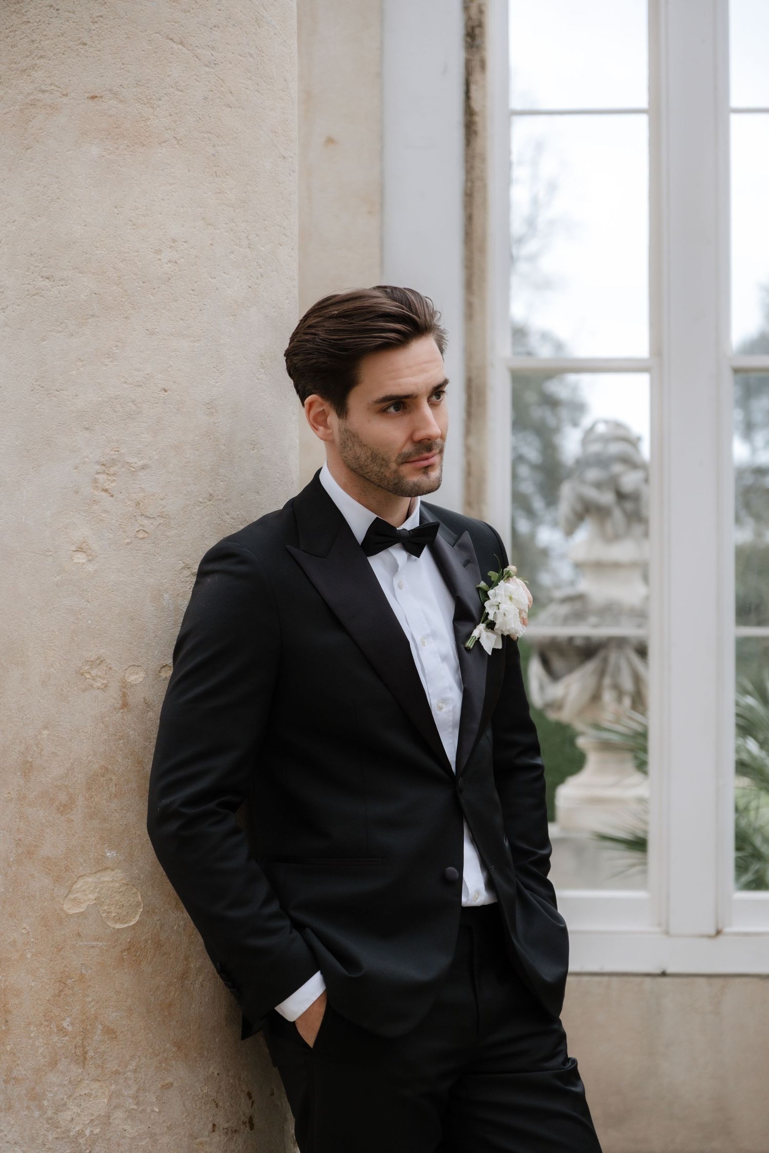 Classic groom portrait in black tuxedo against white conservatory wall.