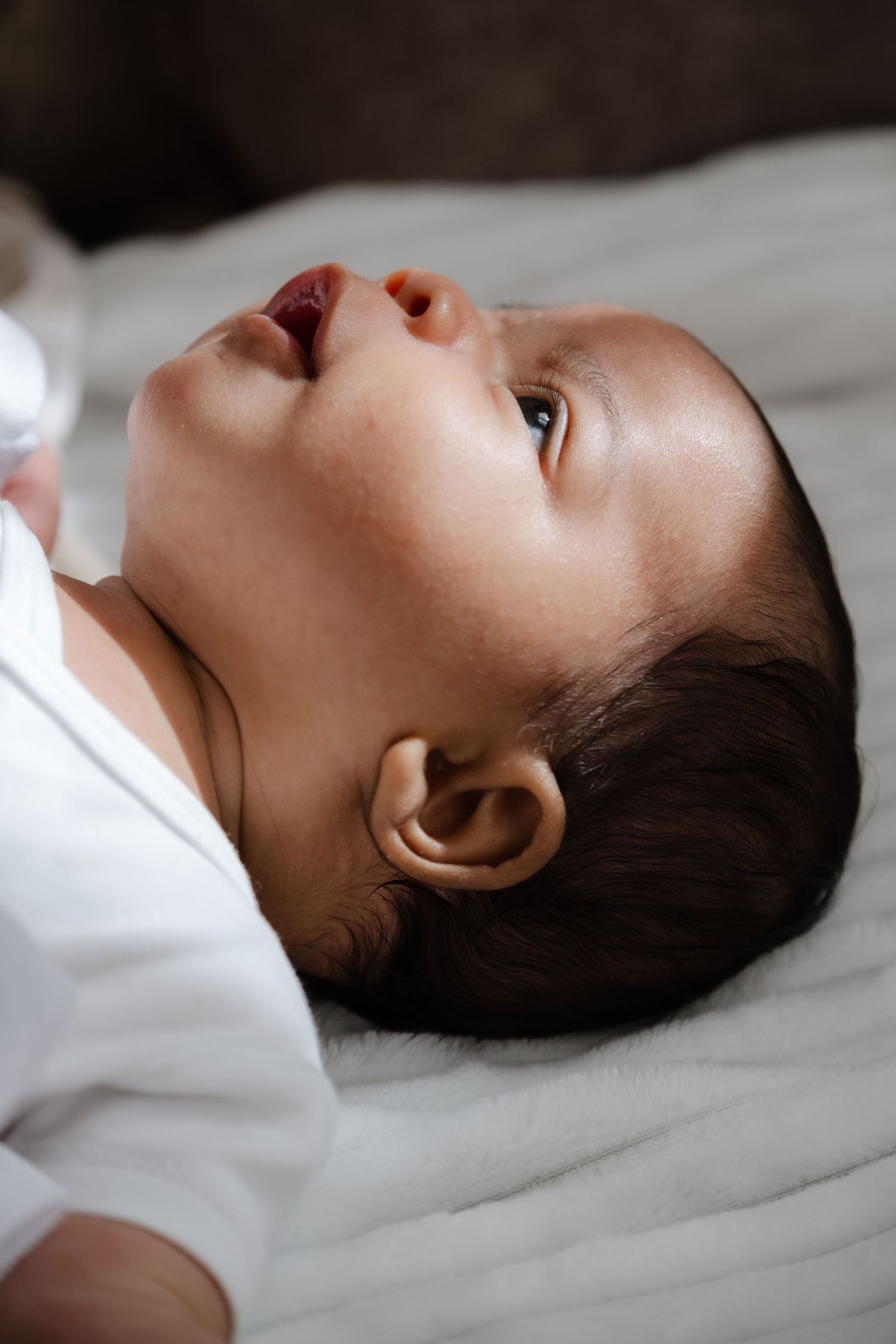 Newborn baby lying on back looking up with mouth open on soft grey blanket.