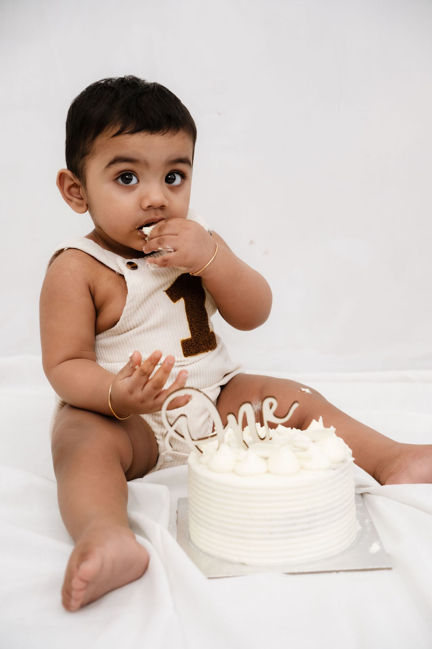 Baby girl celebrates first birthday with white cake smash, wearing cream romper with number one, on white background.