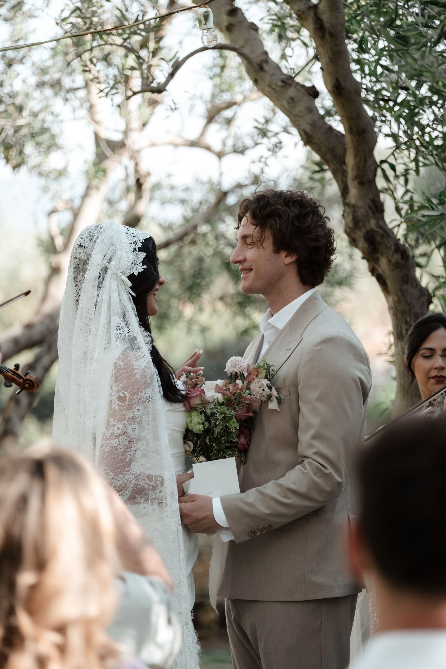 Groom in beige suit smiling at bride in lace veil during outdoor wedding ceremony under olive trees.