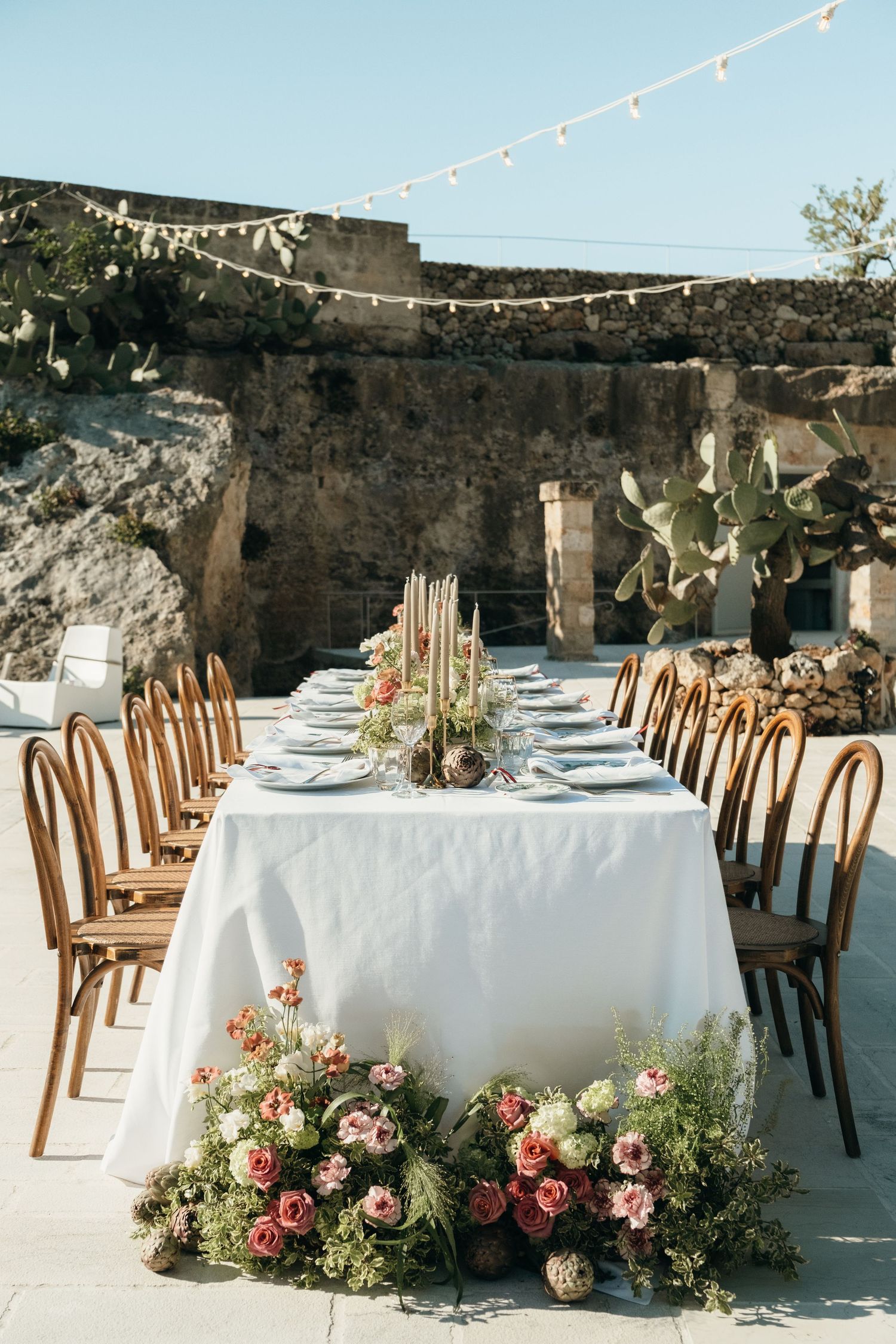 Elegant outdoor wedding reception table with white linen, floral arrangements, candles, and string lights against stone walls.