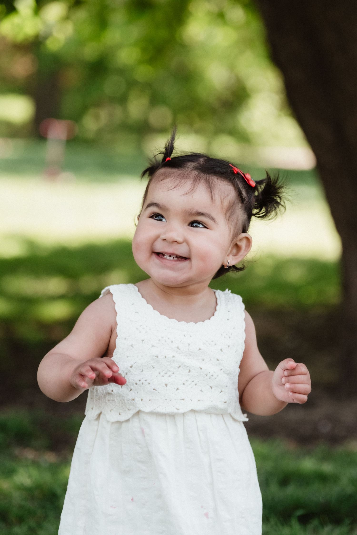 A smiling toddler girl in a white dress with pigtails plays outdoors in a green park on a sunny day.