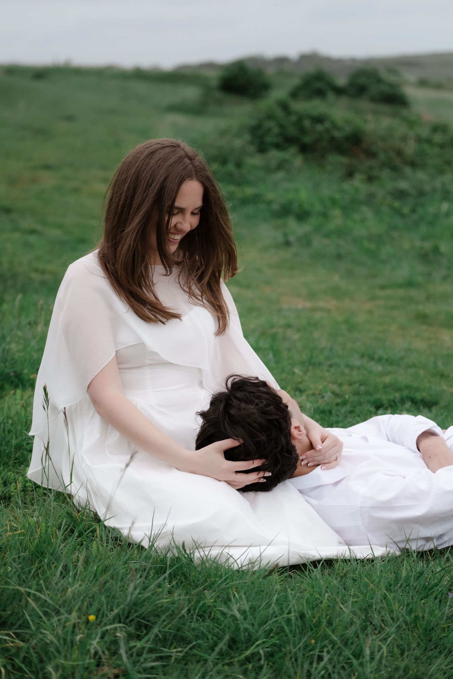 A woman in white dress sits on green grass, smiling down at a person resting their head in her lap.