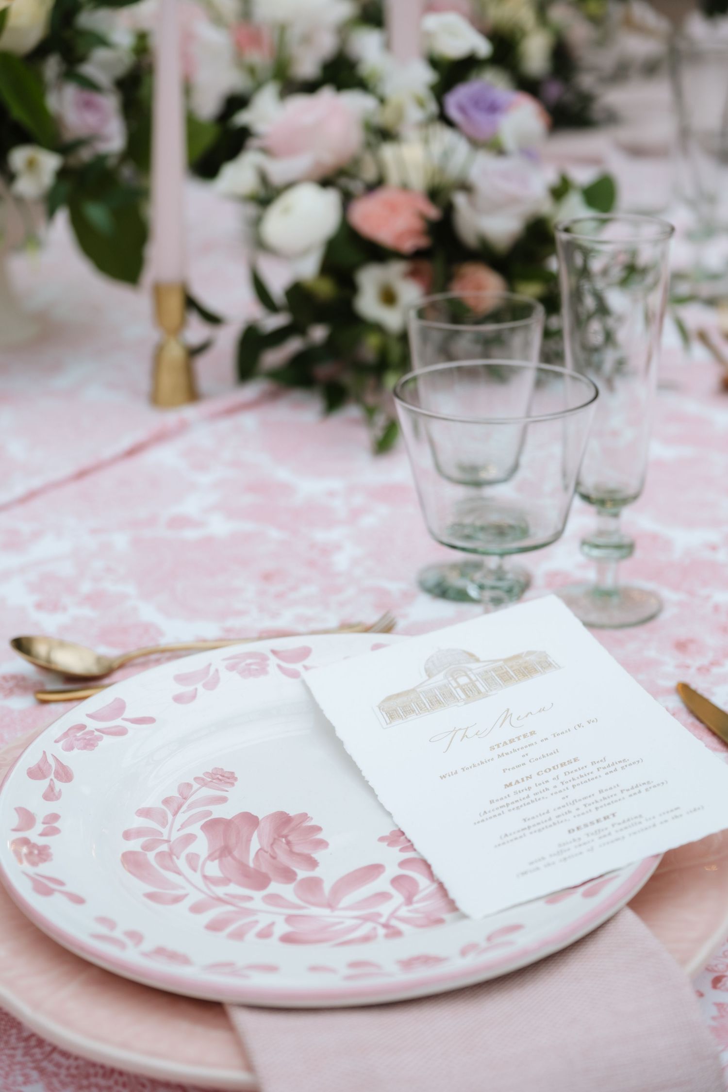 Wedding place setting with pink patterned plates and printed menu cards on white tablecloth.