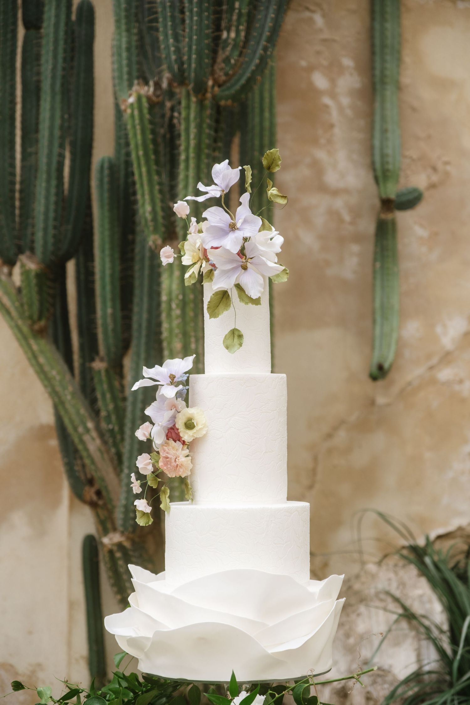 An elegant white tiered wedding cake decorated with delicate purple flowers against a cactus backdrop.