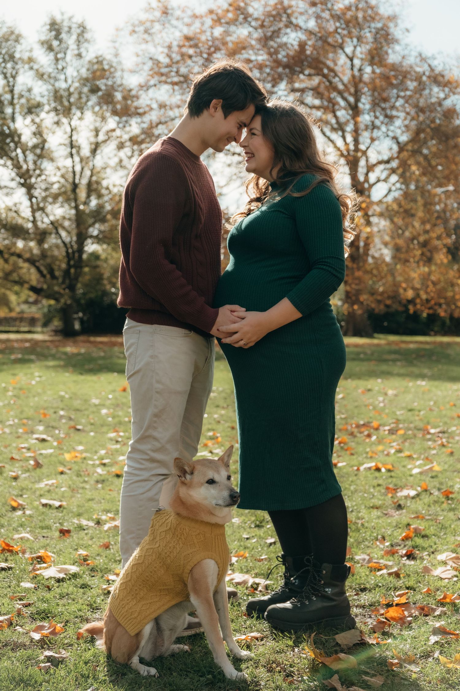 Autumn maternity photoshoot in a park with a dog joining the couple on fallen leaves.