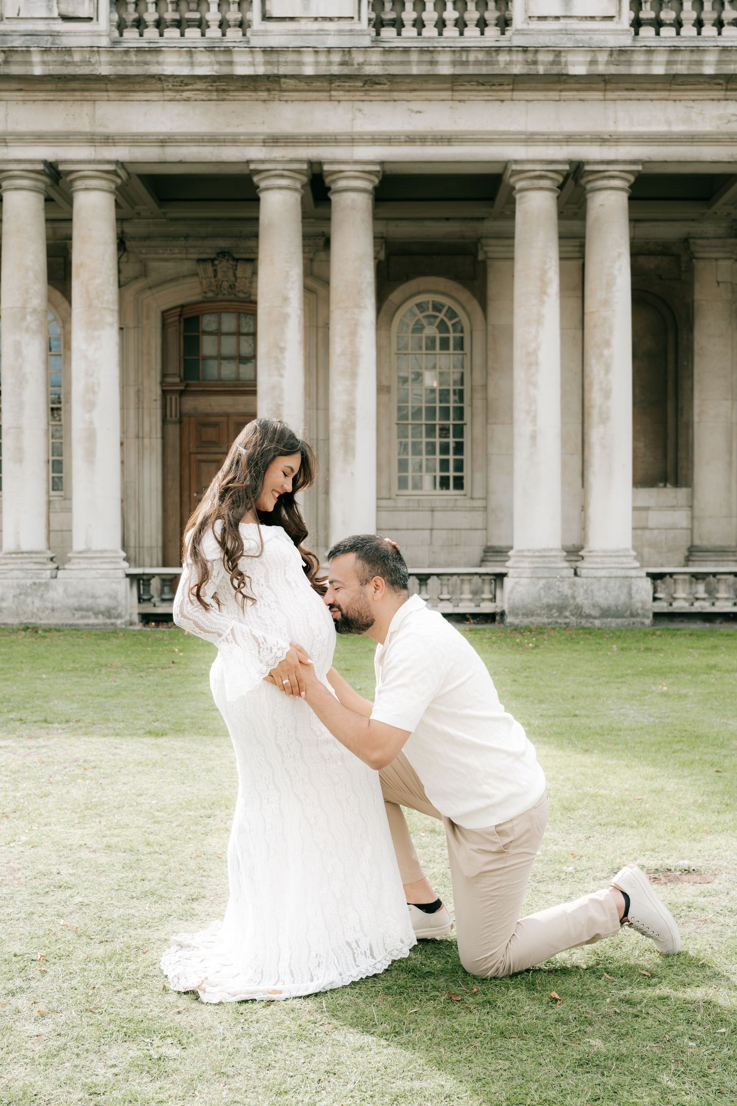 A romantic couple poses playfully on a grassy lawn in front of a classical architectural facade.