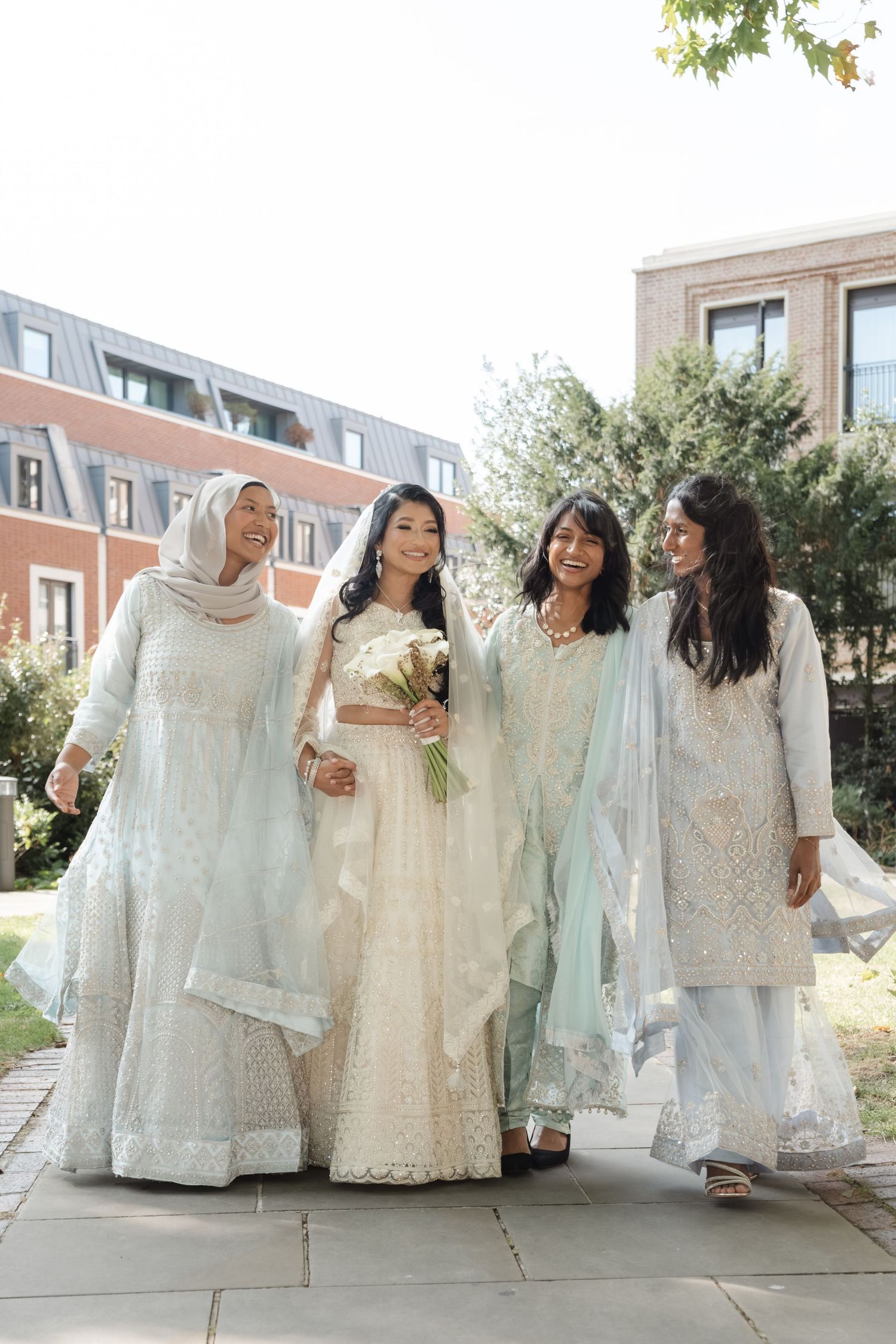 A bridal party in elegant white South Asian outfits walks together on a stone pathway.