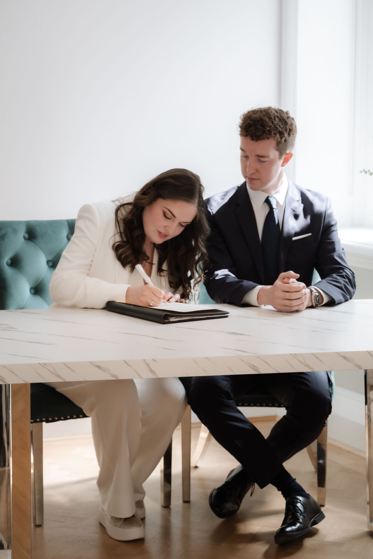 Bride in white suit signing wedding documents at marble table beside groom in black suit.