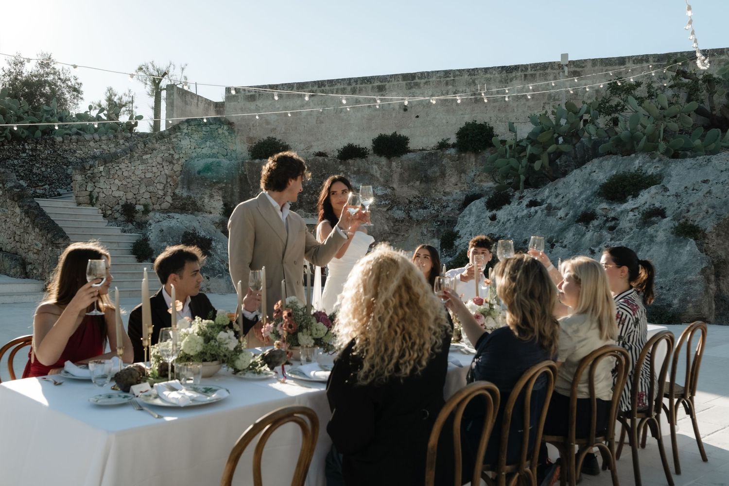 Elegant outdoor dinner party with guests toasting at a beautifully set table against a rustic stone wall backdrop.
