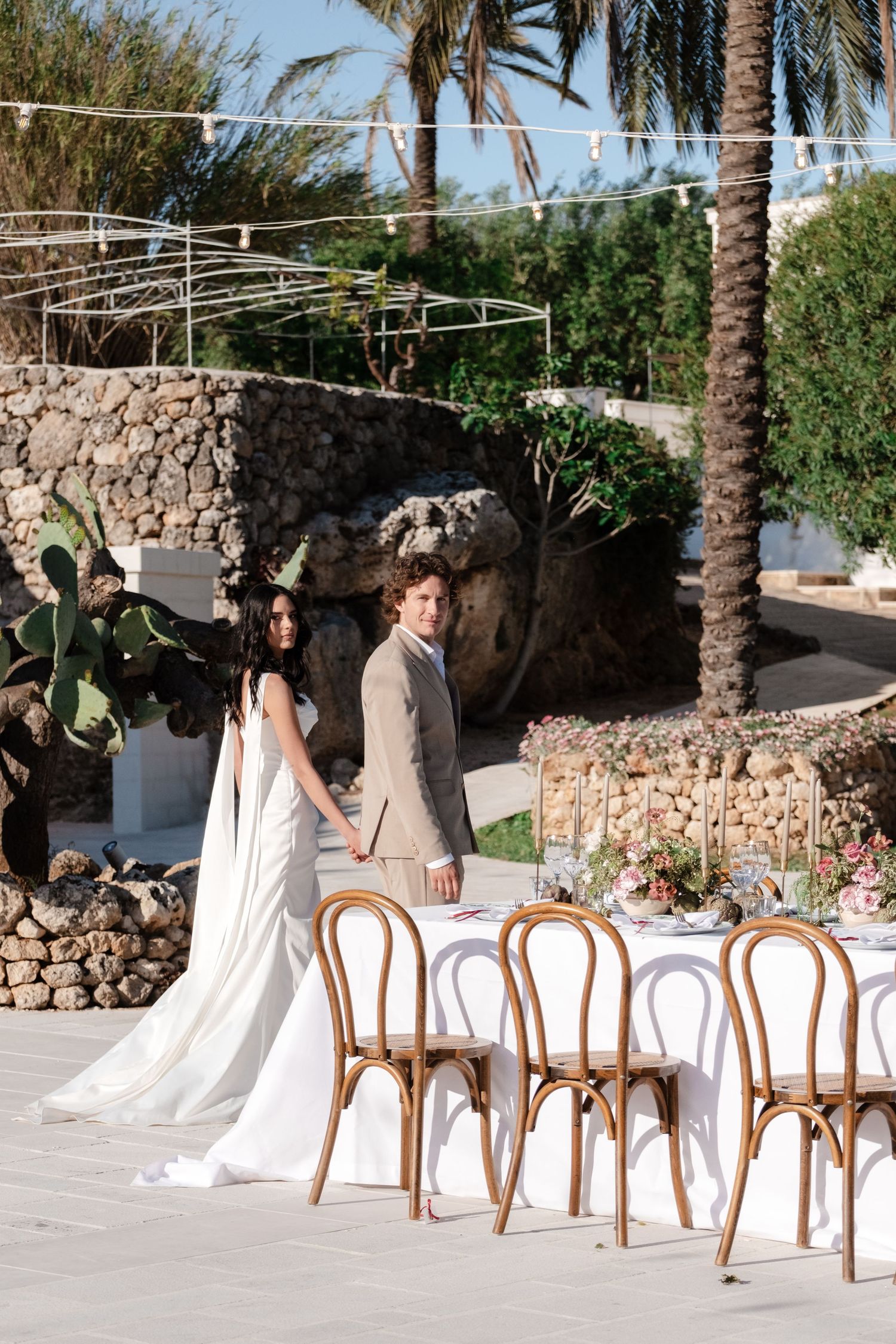 Elegant bride and groom standing beside a decorated outdoor wedding reception table surrounded by palm trees.