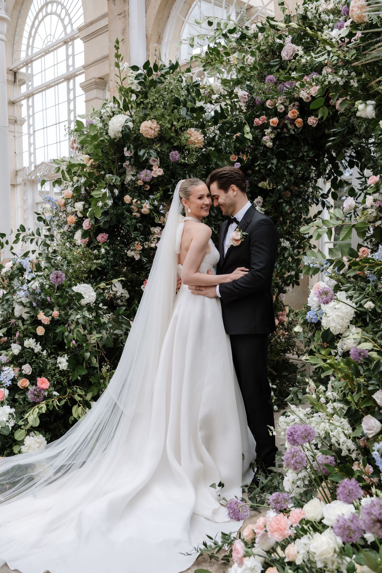 Elegant wedding scene with floral arches and white gown in a sunlit conservatory.