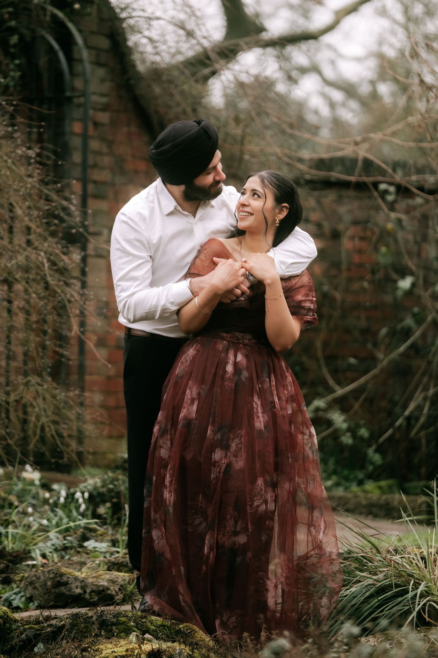 A romantic couple embraces in a garden setting, with her wearing a flowing burgundy dress against brick walls.