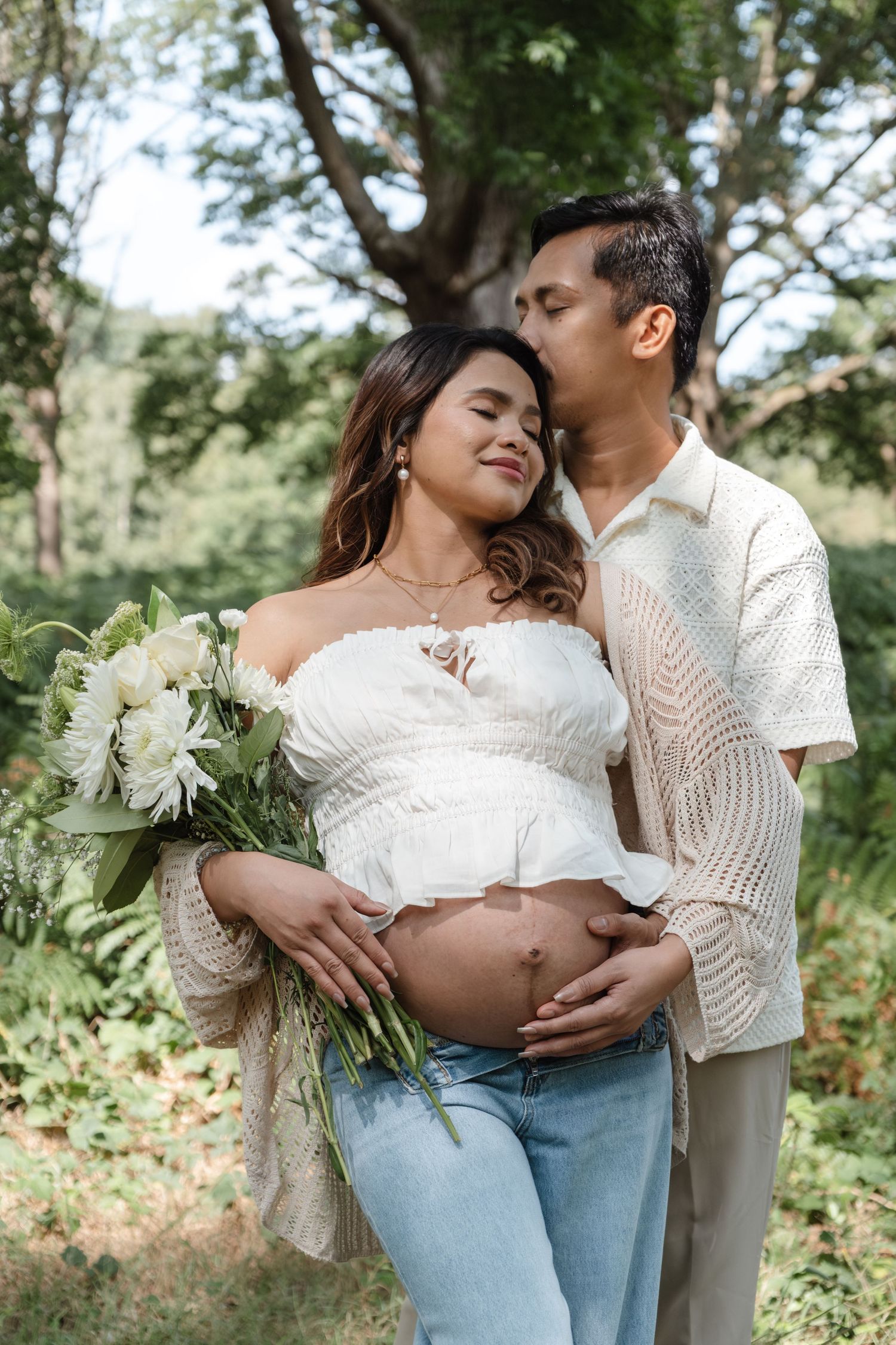 Casual maternity photos with couple in white tops and jeans holding flowers outdoors.