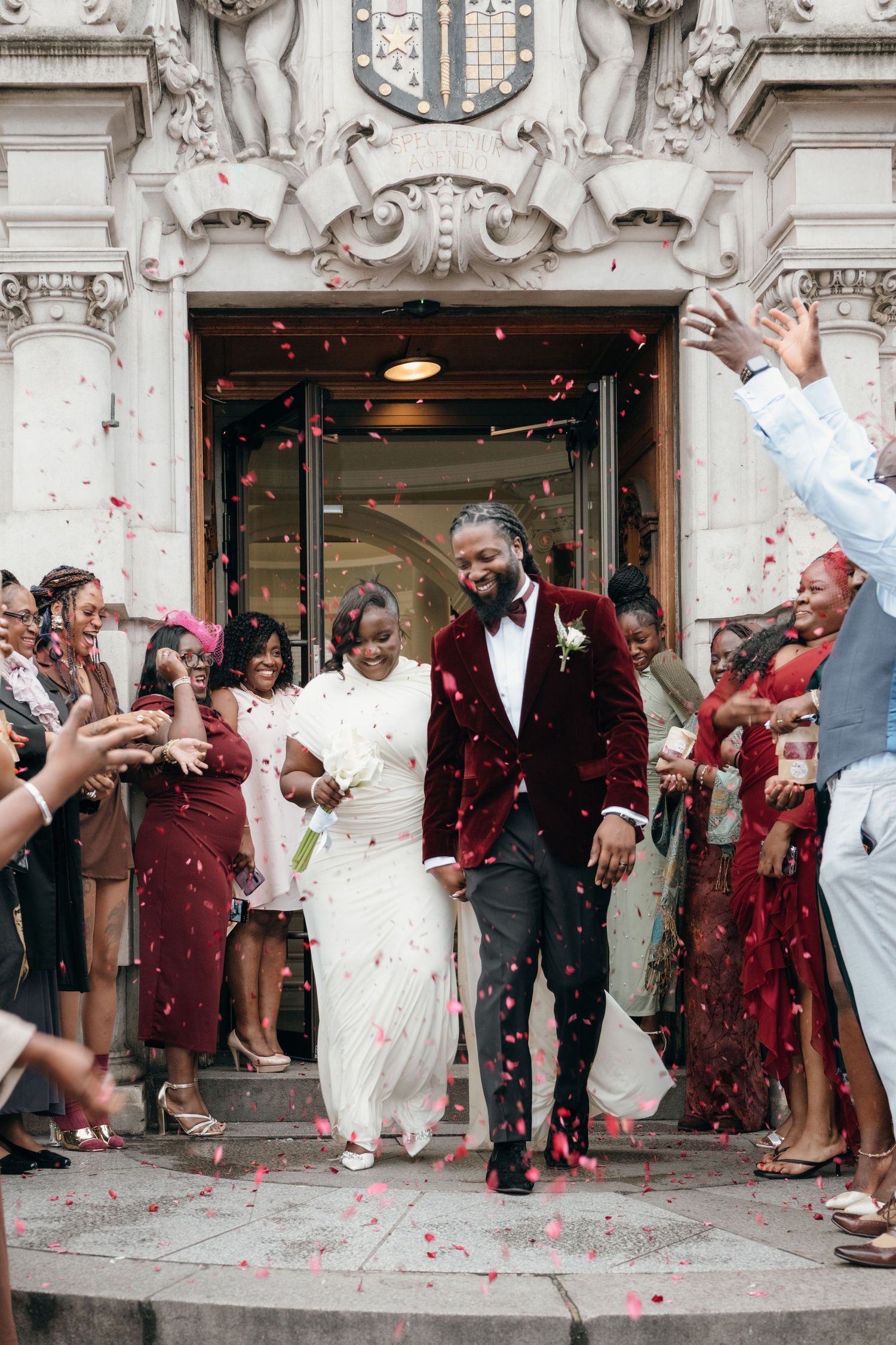 Wedding guests throw confetti as newlyweds exit an ornate building entrance.