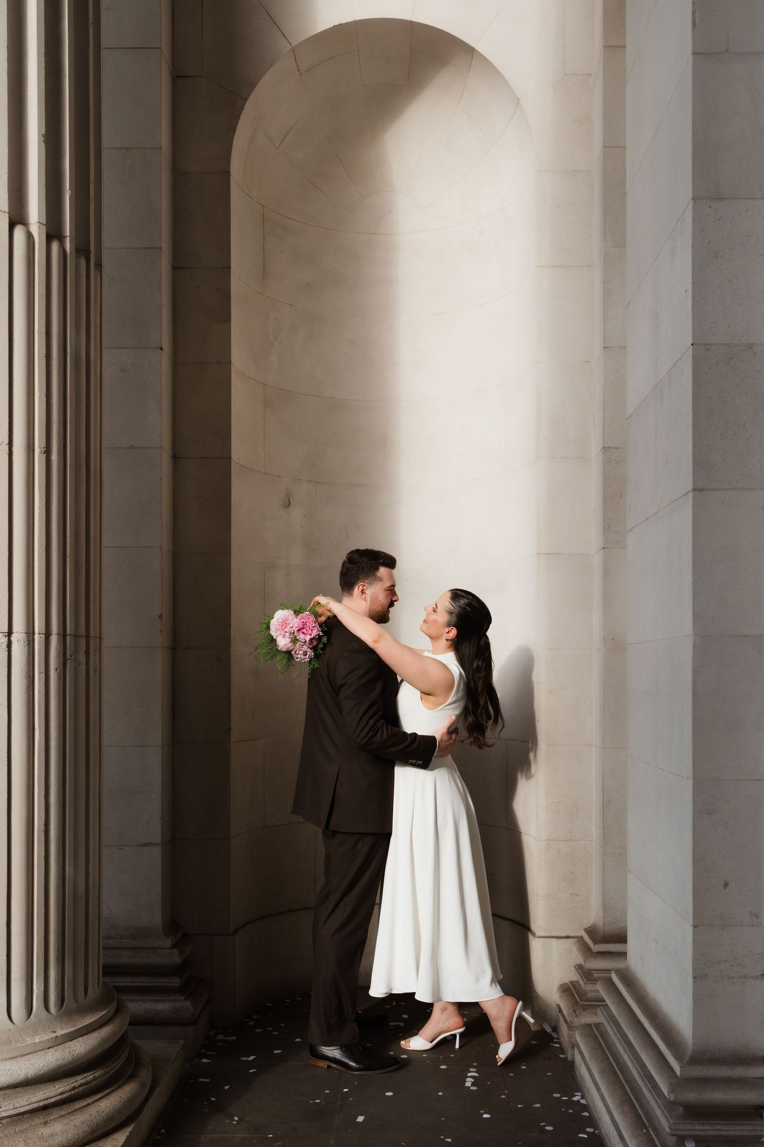Bride and groom embracing in an elegant arched stone colonnade, bride holding pink bouquet in white dress.
