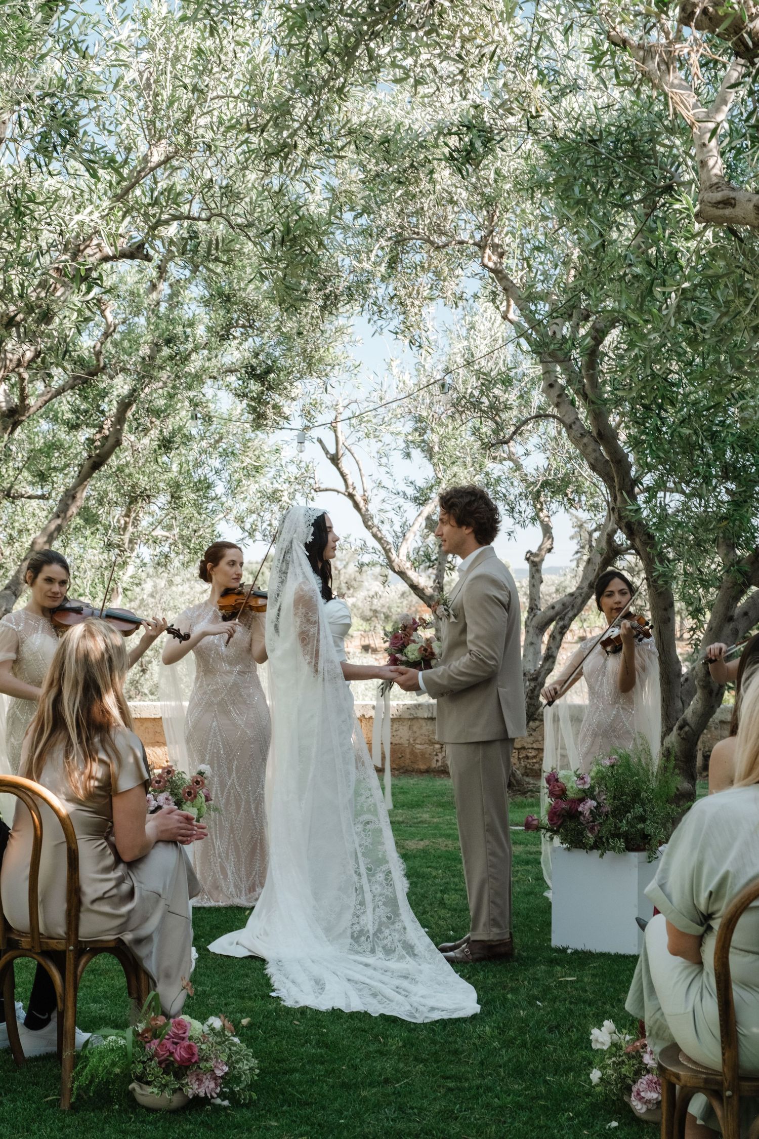 Outdoor wedding ceremony under olive trees with violinists, bride in lace gown, groom in tan suit, floral decor.
