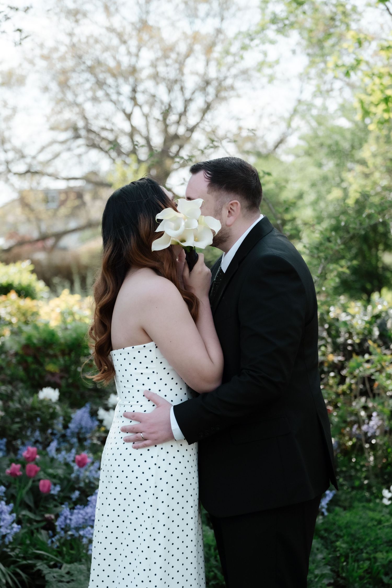 Couple sharing a kiss hidden behind white flowers in a lush garden, wearing formal attire for a wedding.