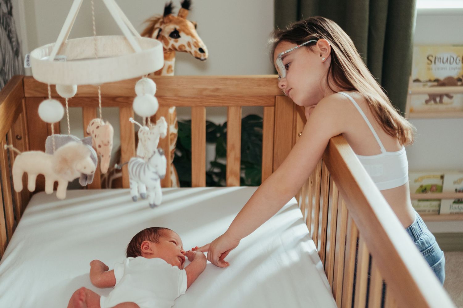 Young girl gently touching a newborn baby lying in a wooden crib with animal mobile above.