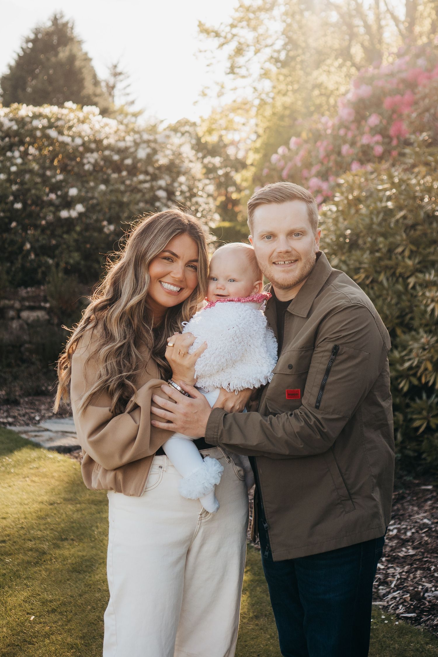 Smiling family with mother, father, and baby posing together in a sunny blooming garden.