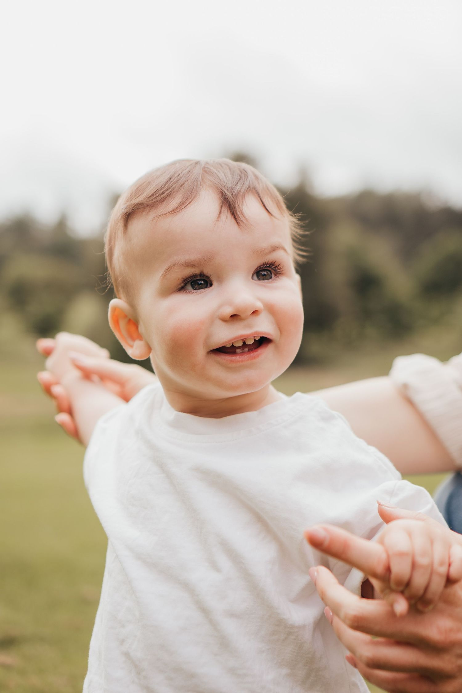 Happy toddler in white t-shirt smiling outdoors, held by mum hands during his first birthday milestone session