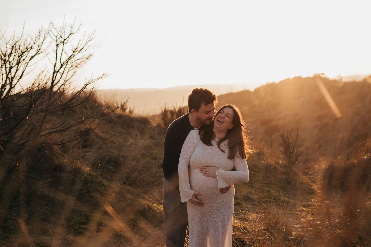 Couple embracing at golden sunset in a field, pregnant woman in white dress held tenderly by partner outdoors.