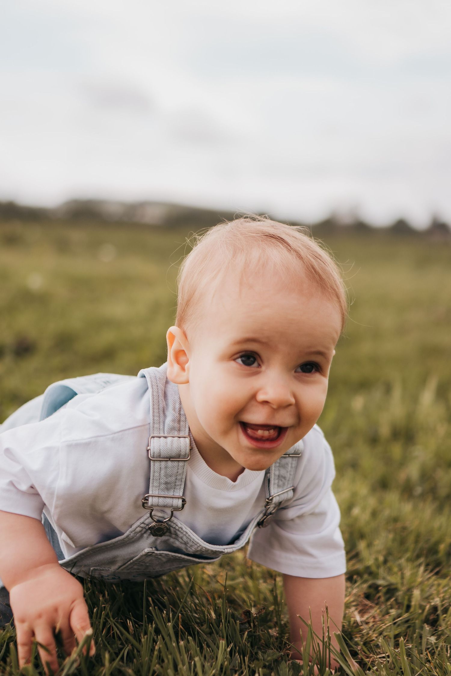 A cheerful baby in denim overalls crawling on green grass outdoors, smiling at the camera on a cloudy day during his first birthday session