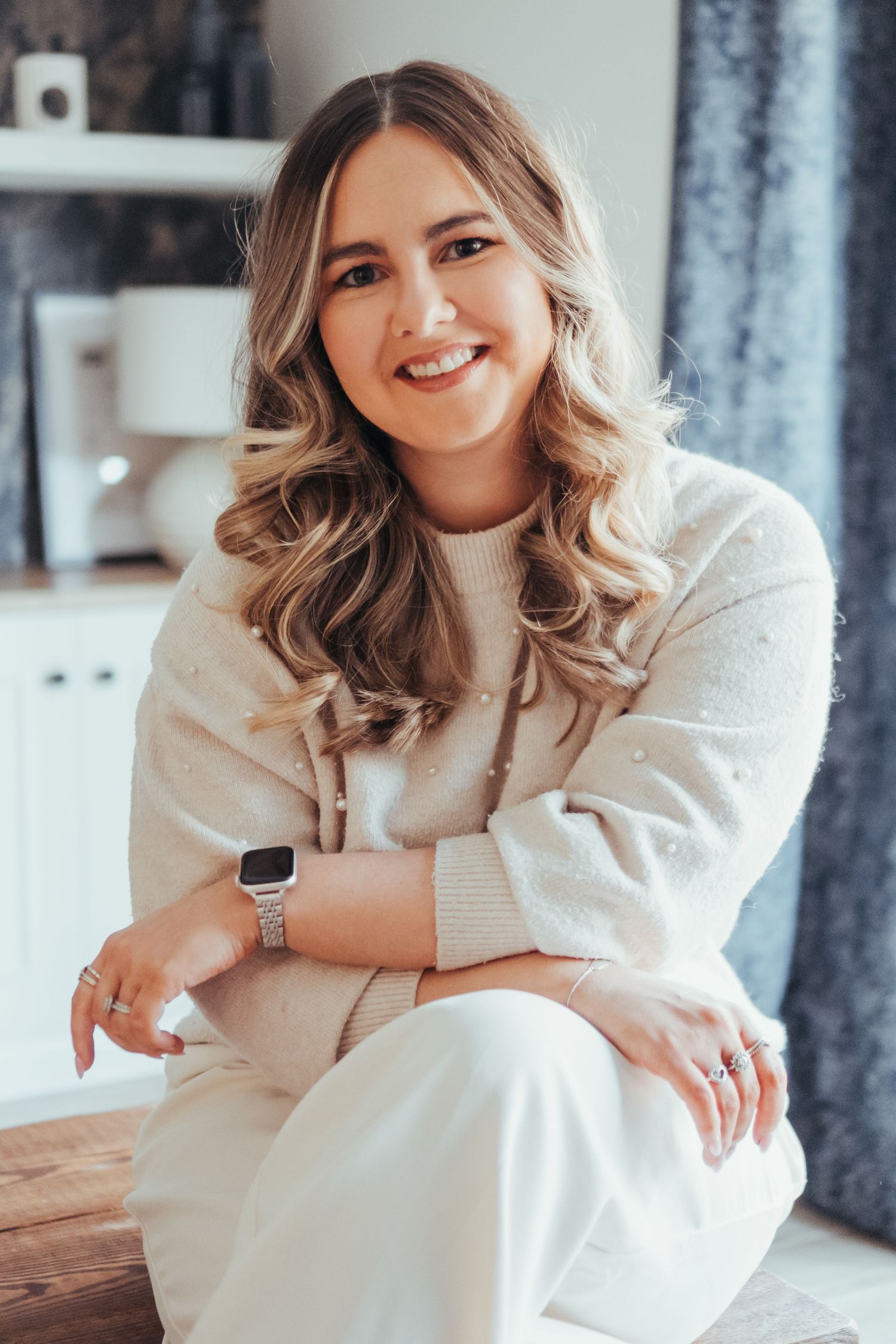 Happy young woman with wavy blonde hair wearing a cream knit sweater and smartwatch, sitting casually indoors.
