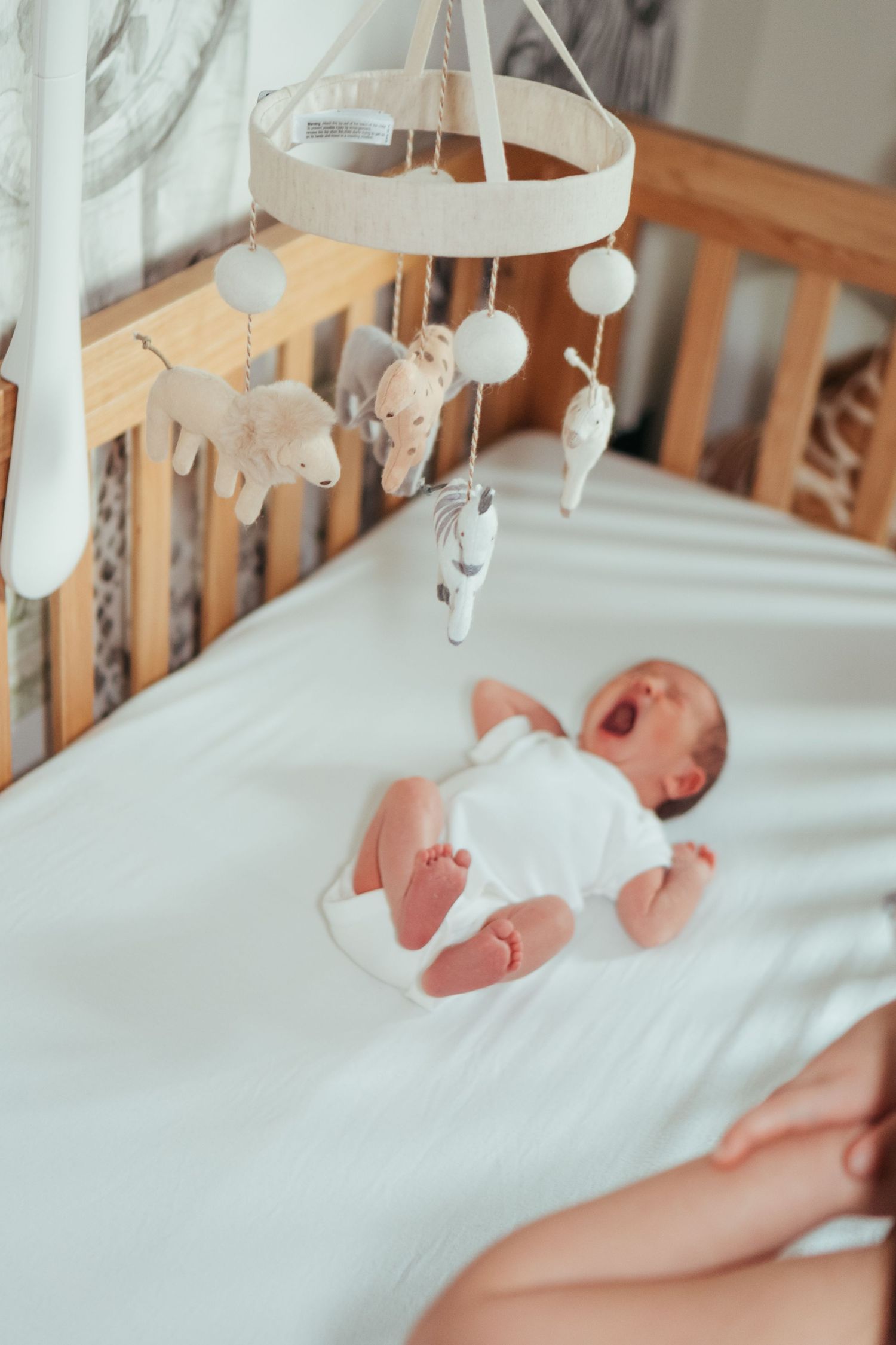 Newborn baby yawning in a white onesie inside a wooden crib with a hanging animal mobile.