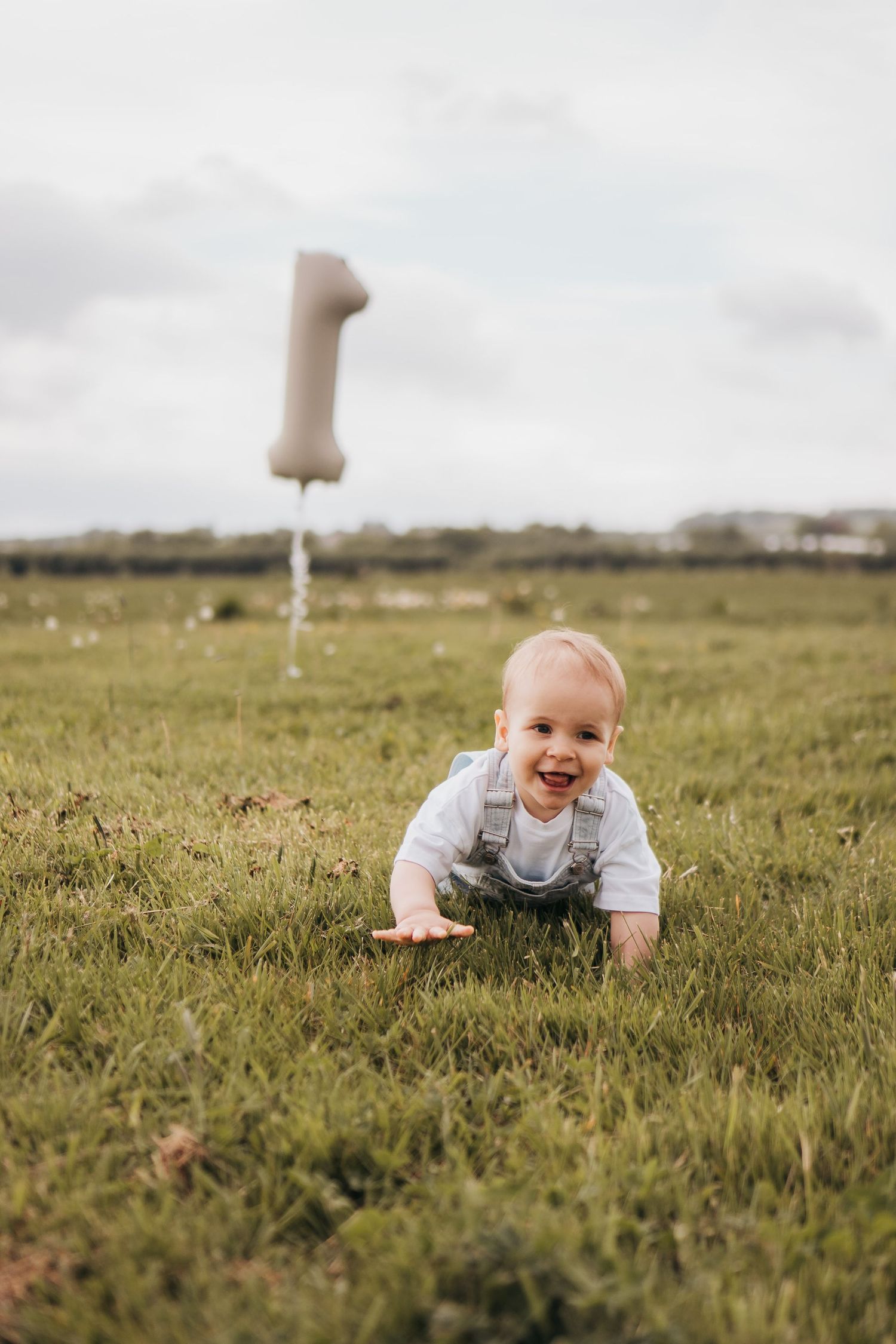 Happy baby boy crawling on green grass outdoors with a number one balloon in the background.
