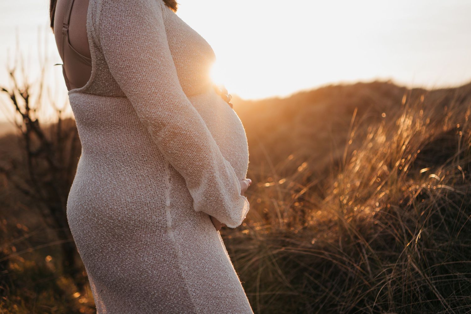 Pregnant woman in beige dress holding baby bump at golden sunset in nature field.