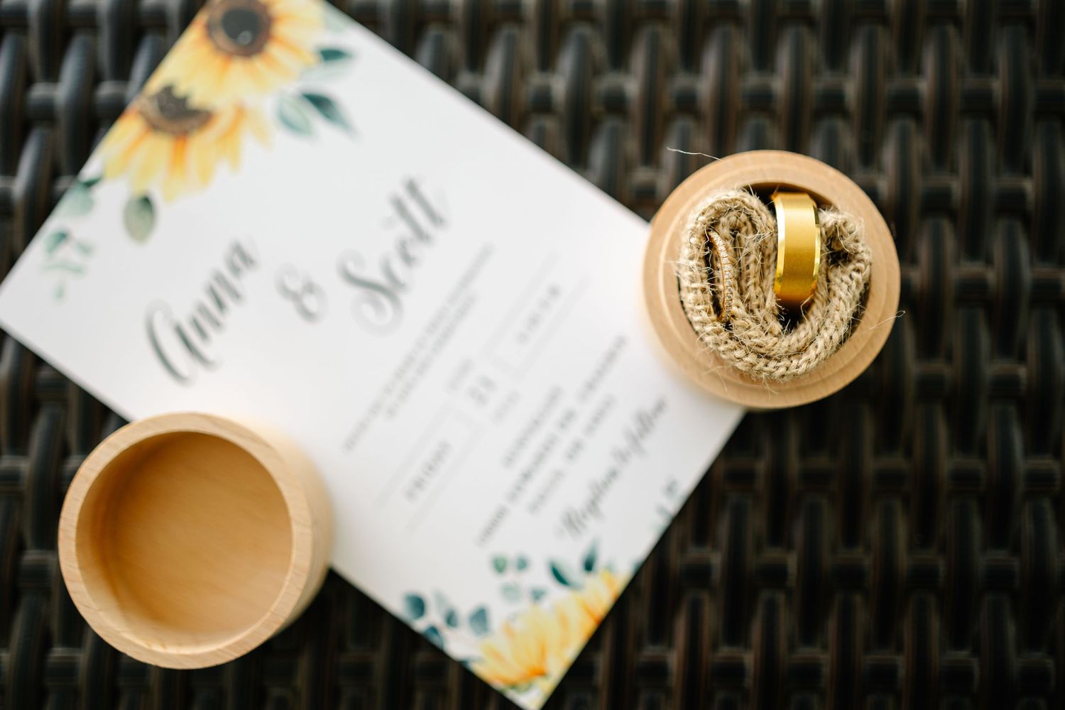 Wedding rings displayed next to a floral invitation card on a dark woven surface.