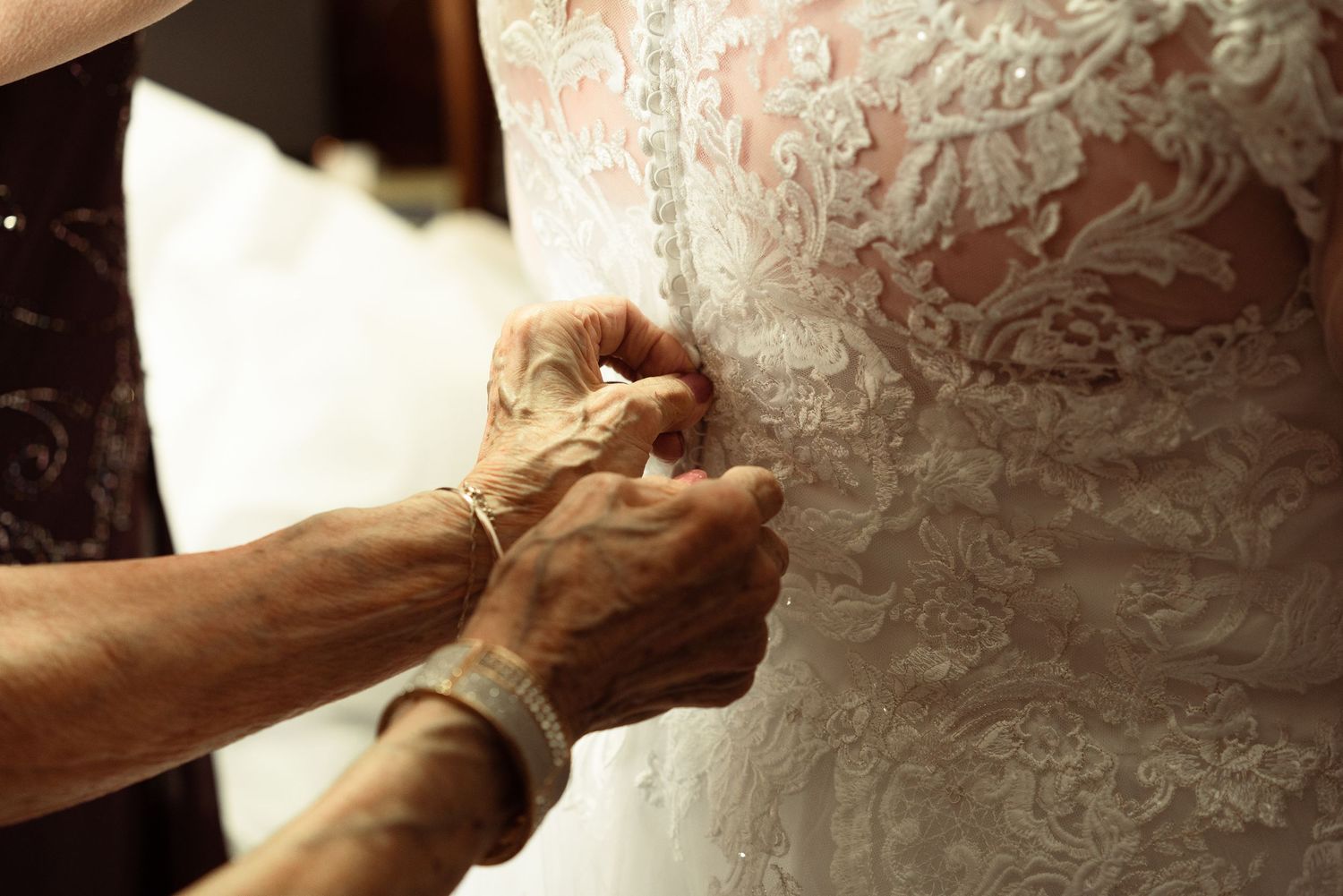 Close-up showing hands buttoning up the back of an ornate white lace wedding dress.