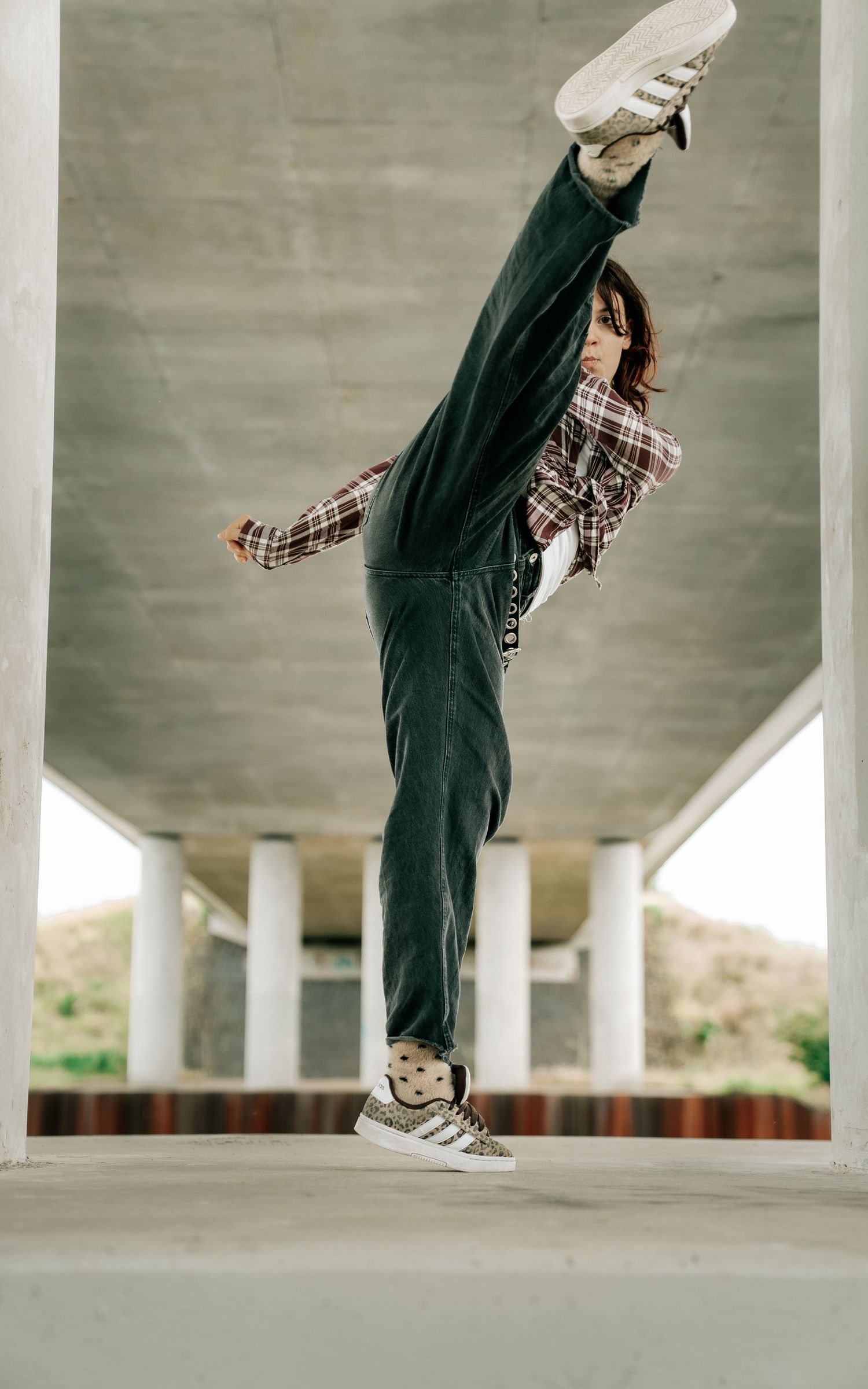 Young woman performing high kick in plaid shirt and dark jeans under concrete bridge.