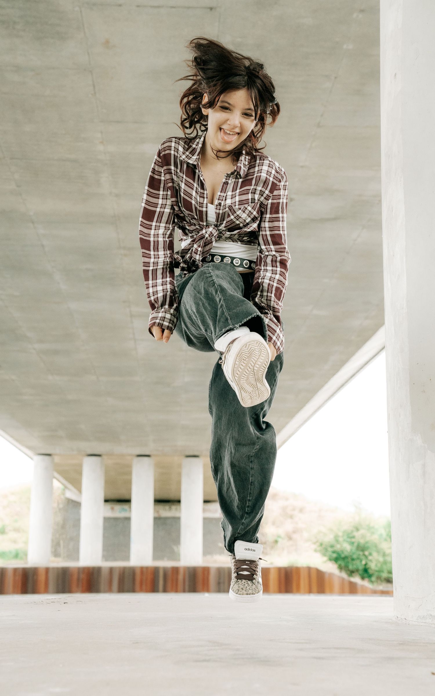 Young girl in plaid shirt and baggy jeans jumping mid-air under a concrete overpass, smiling joyfully.