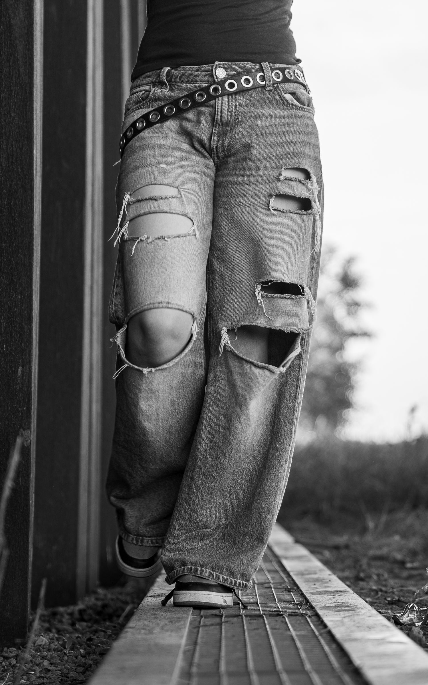 Black and white photo of ripped baggy jeans with a studded belt, worn with sneakers on a wooden walkway.
