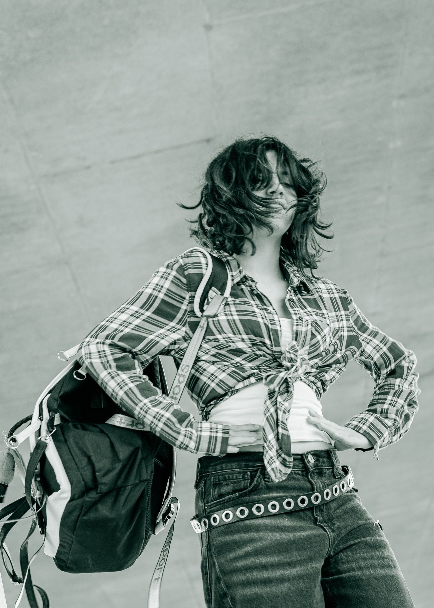 Woman in plaid shirt and jeans with backpack posing outdoors, hair blowing in wind, black and white photo.