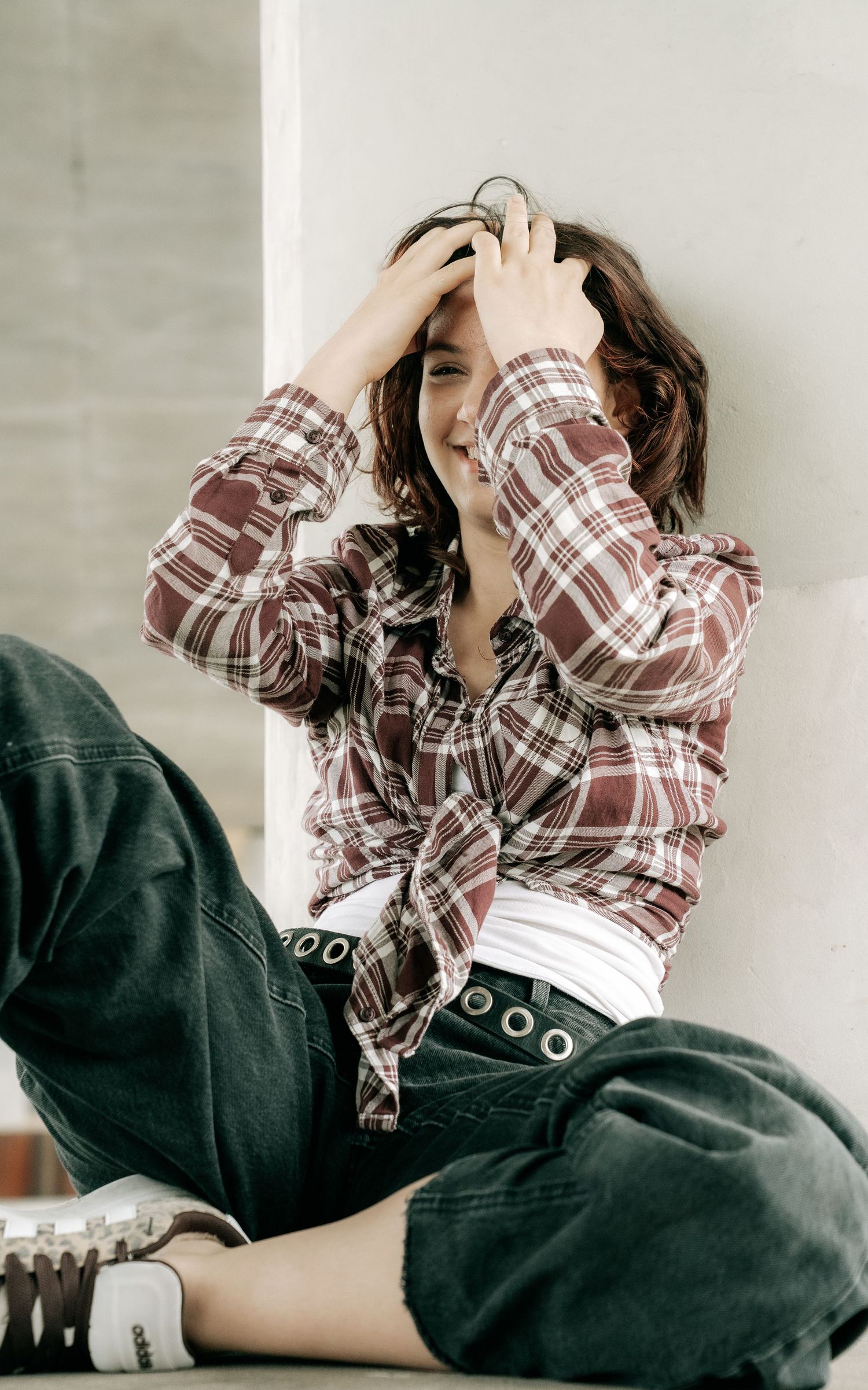 Young woman in plaid shirt and dark jeans sitting against wall laughing with hands in hair.