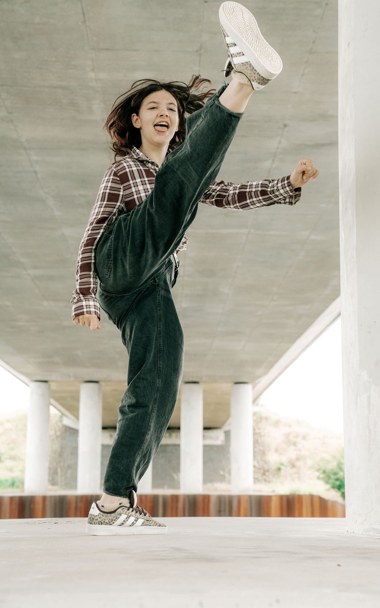 A young woman joyfully kicks her leg high while dancing under a concrete overpass, wearing overalls and a plaid shirt.