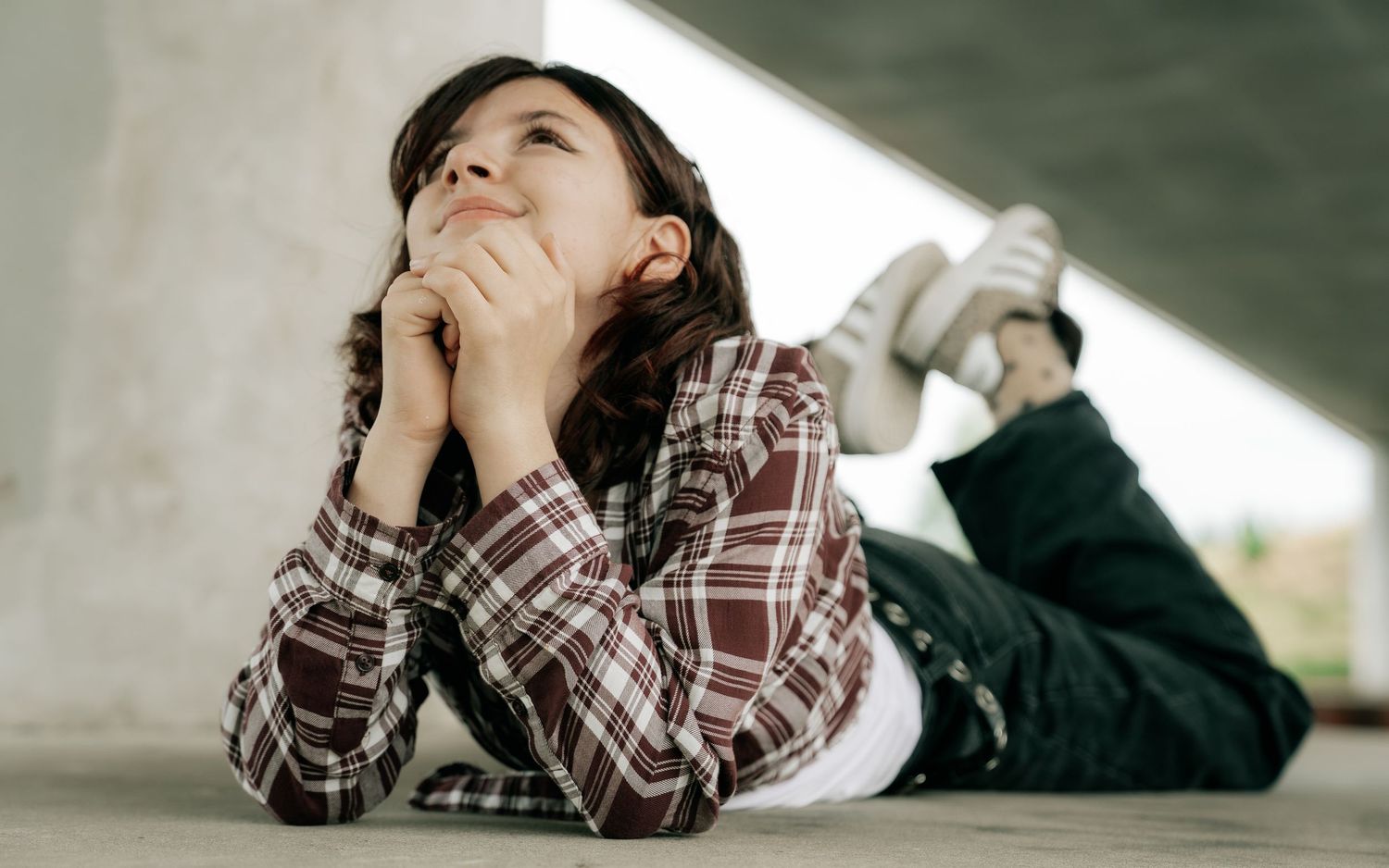 Young girl lying on ground under bridge, resting chin on hands, gazing upward thoughtfully in casual plaid shirt.