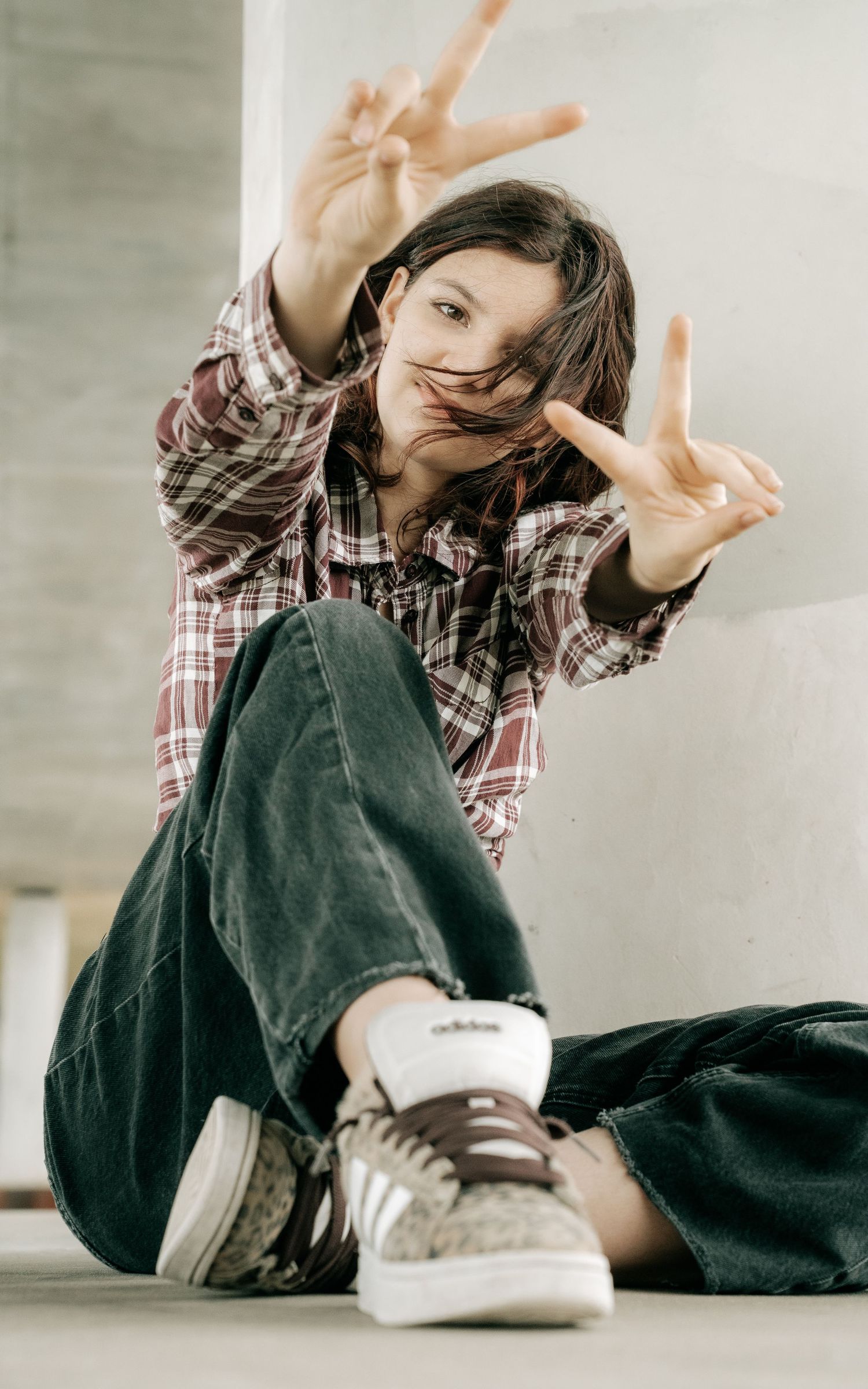 A young person sitting on the ground making peace signs, wearing a plaid shirt, jeans, and Adidas sneakers.