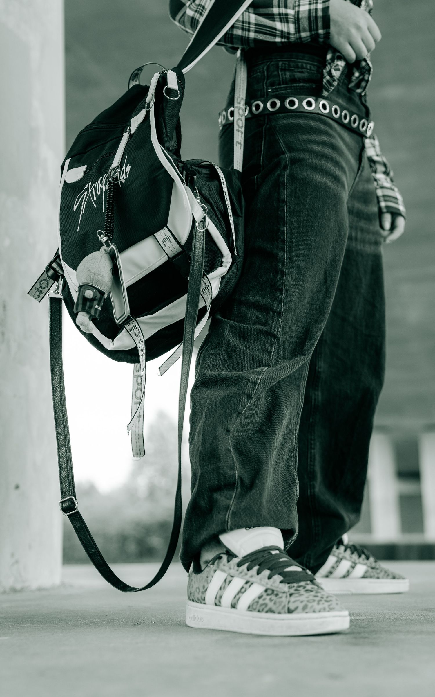 Person holding a backpack wearing jeans, plaid shirt, studded belt, and Adidas sneakers in a black and white photo.