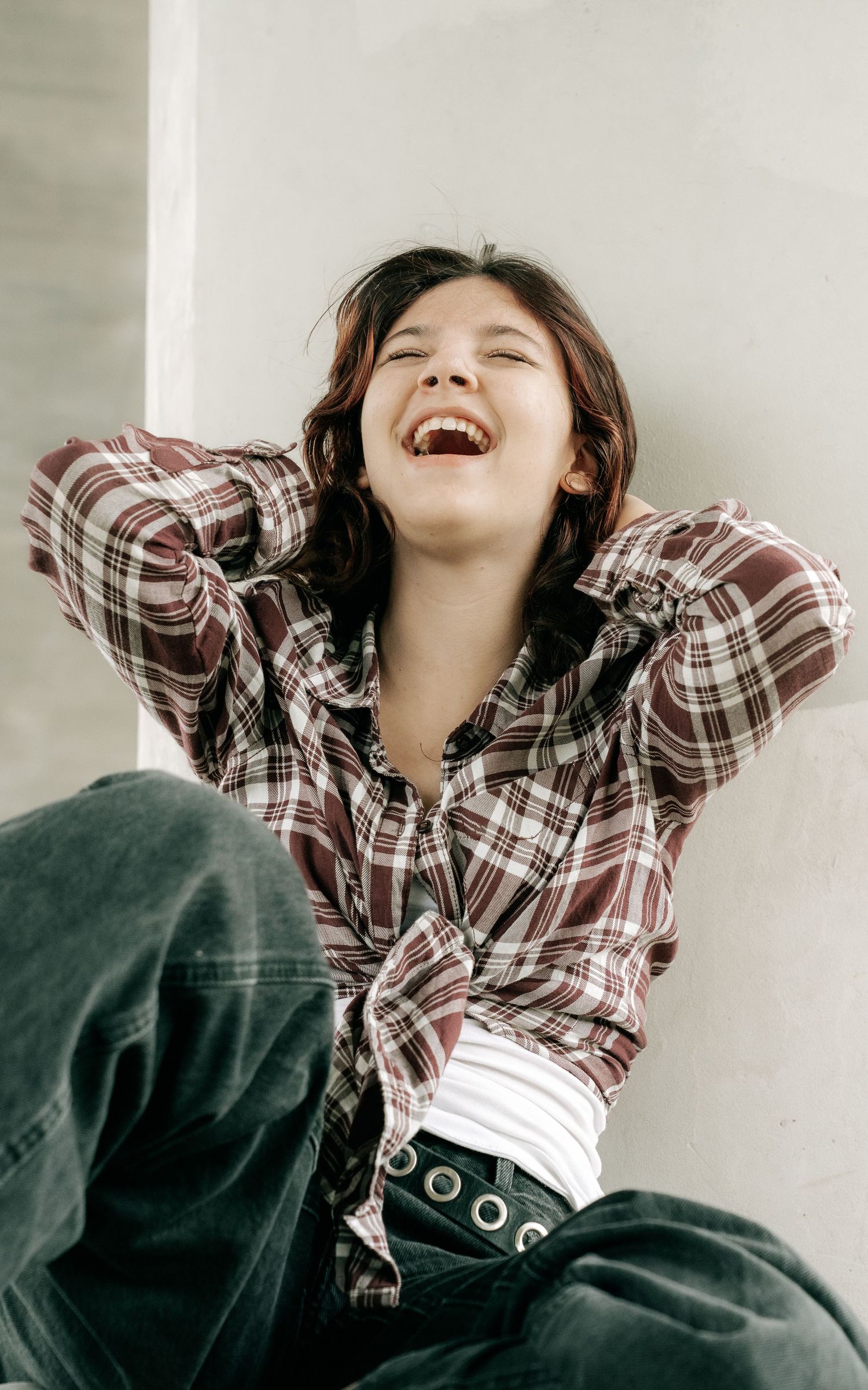 A young woman laughs joyfully while leaning against a wall in a plaid shirt and dark jeans.