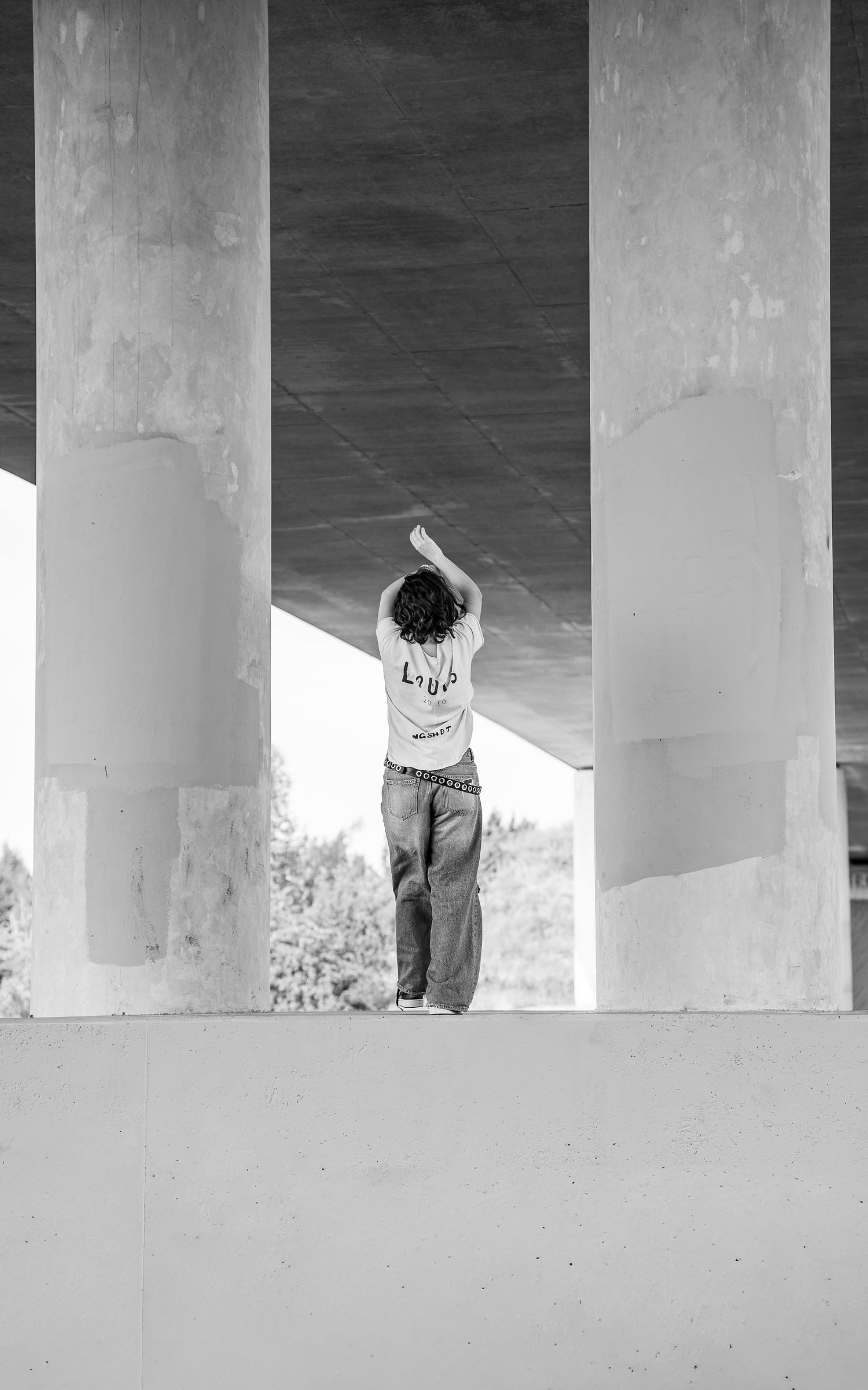Black and white photo of a person standing between concrete pillars under a bridge with arms raised overhead.