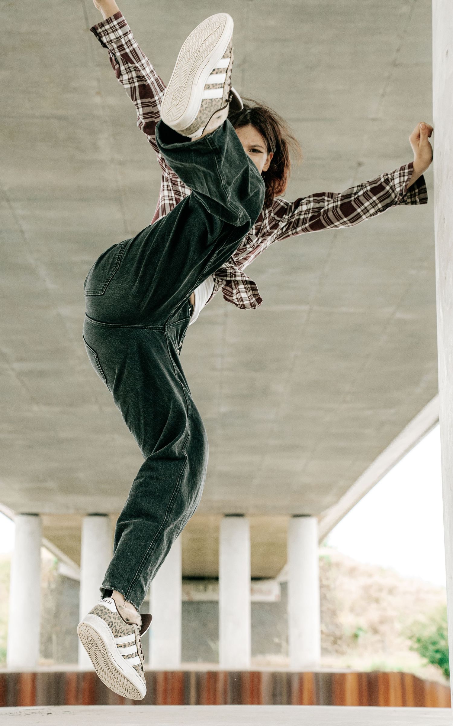 A young dancer leaps mid-air under a concrete bridge, wearing green overalls and a plaid shirt with white sneakers.