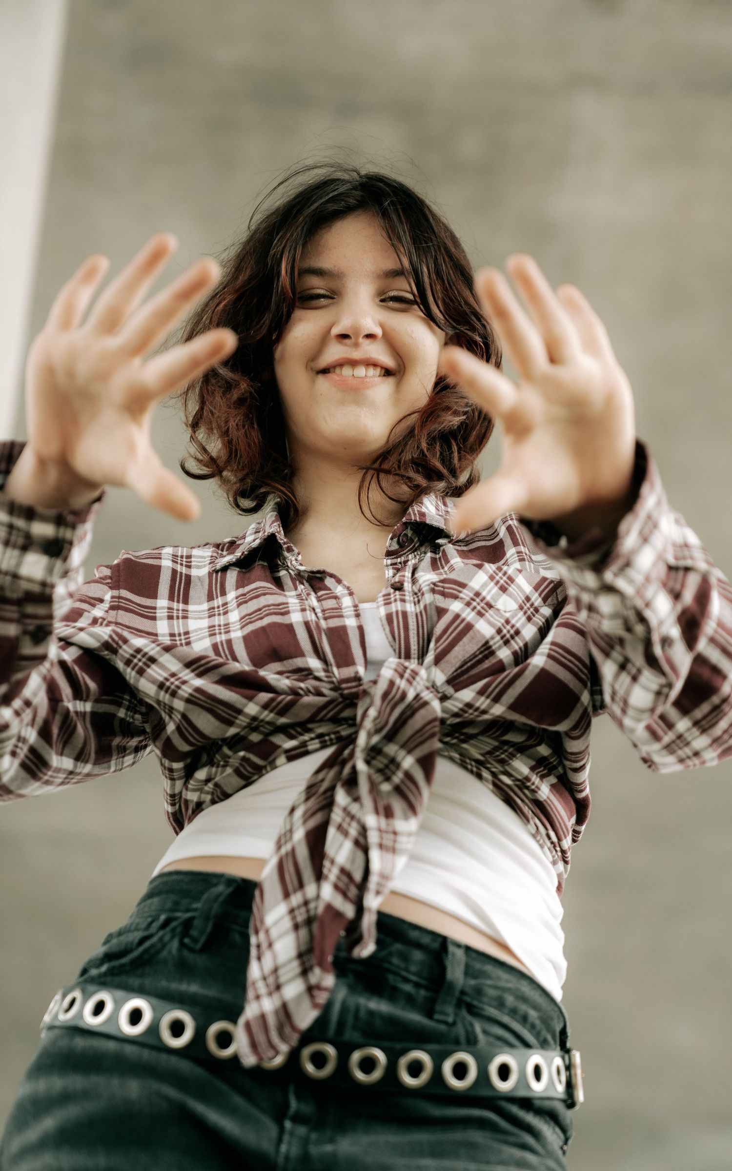 A smiling young woman in a tied plaid shirt reaches her hands toward the camera playfully.