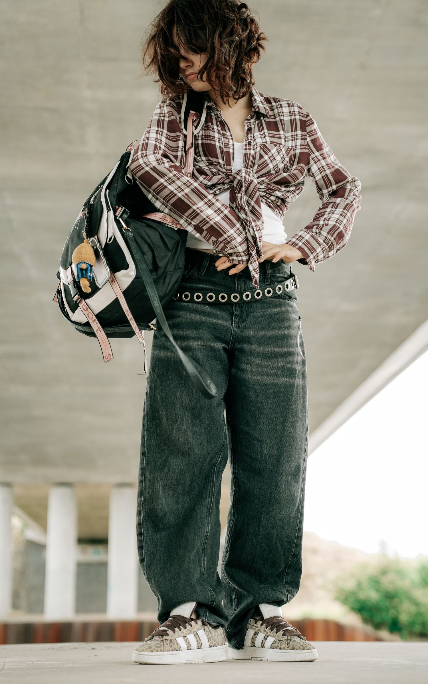 A stylish young woman poses in a tied plaid shirt, wide-leg jeans, and sneakers with a backpack.
