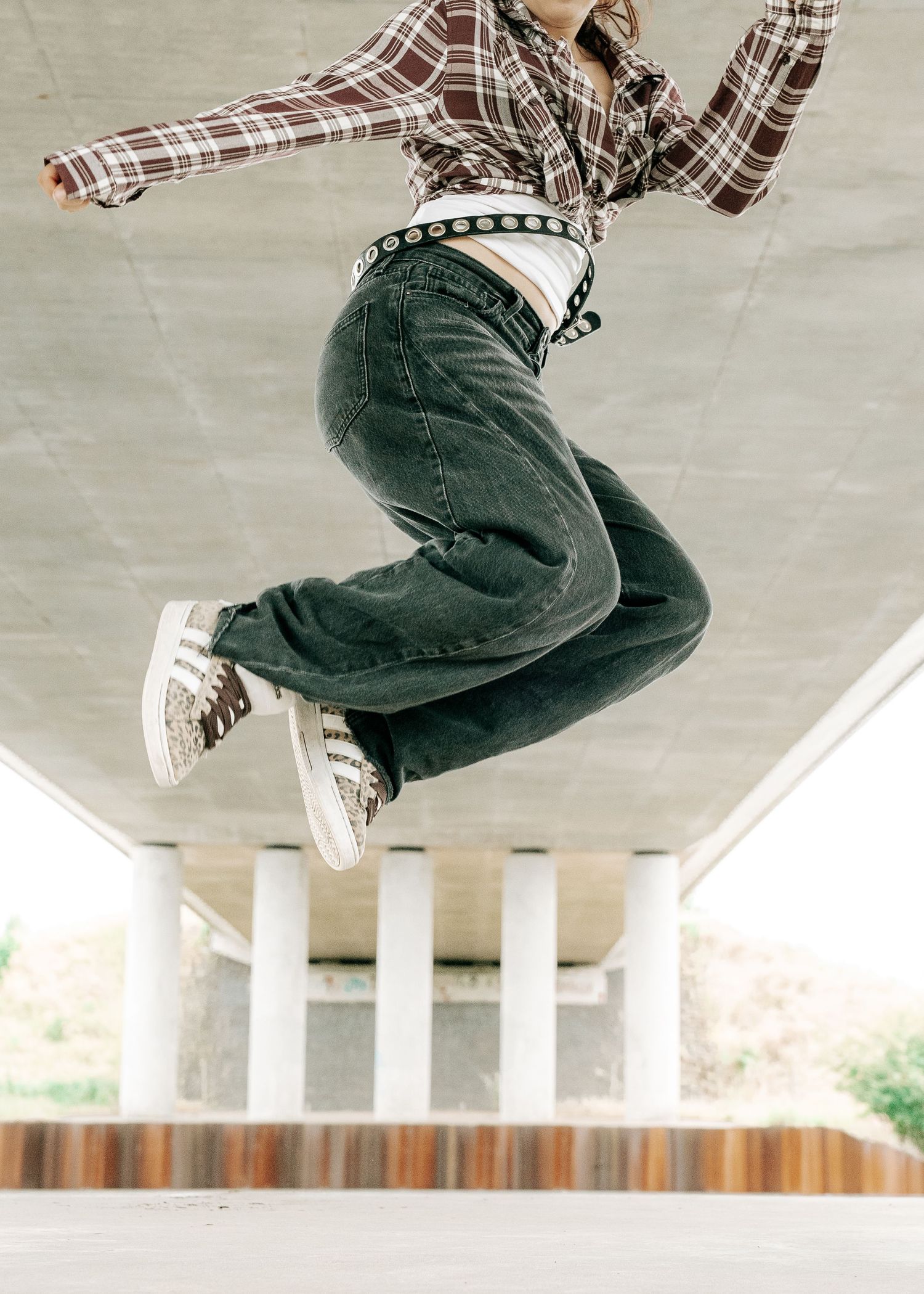 Young woman in plaid shirt and dark jeans jumping energetically under a concrete overpass wearing sneakers.