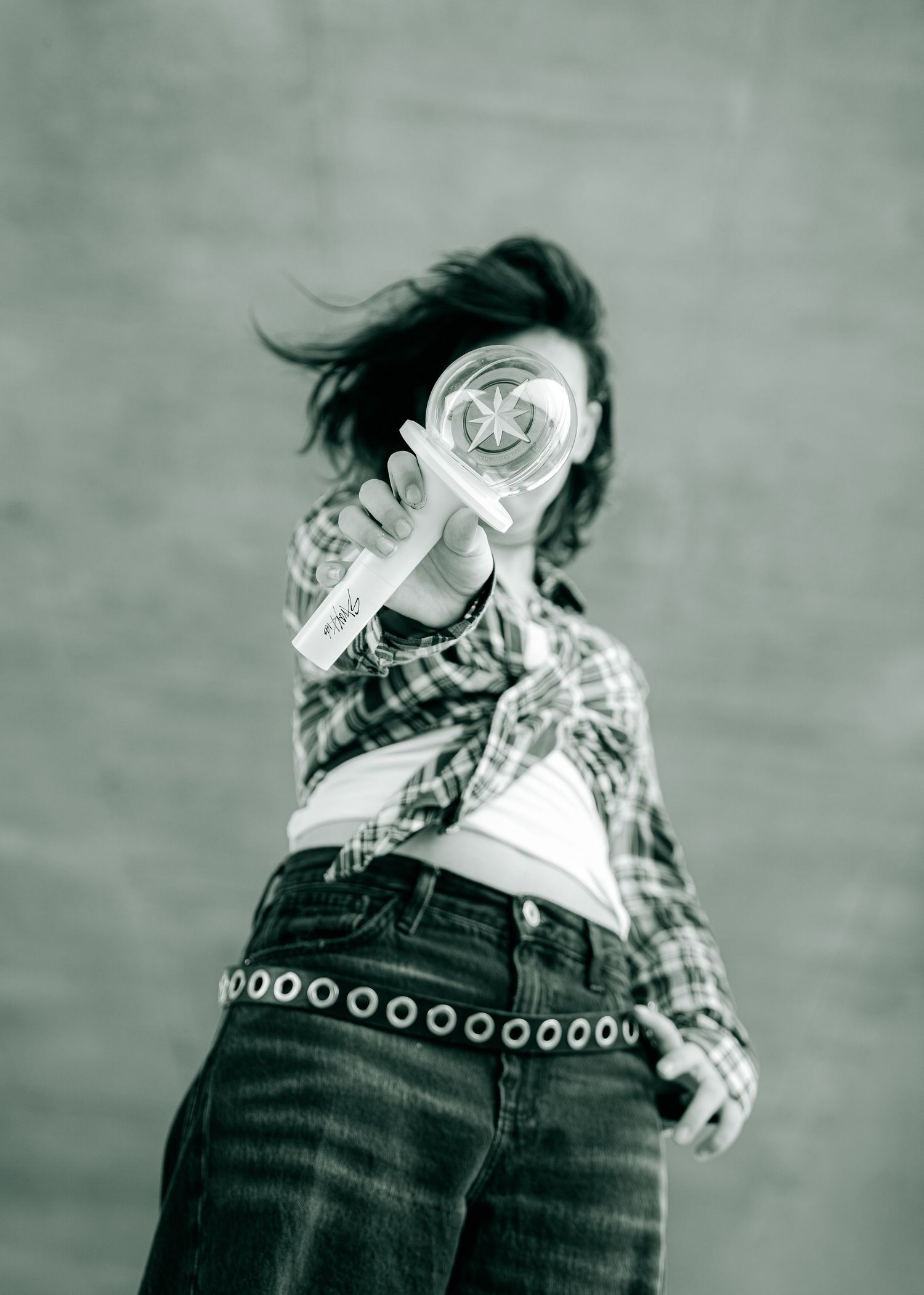 Black and white photo of a woman in plaid shirt and jeans holding a lollipop in front of her face outdoors.